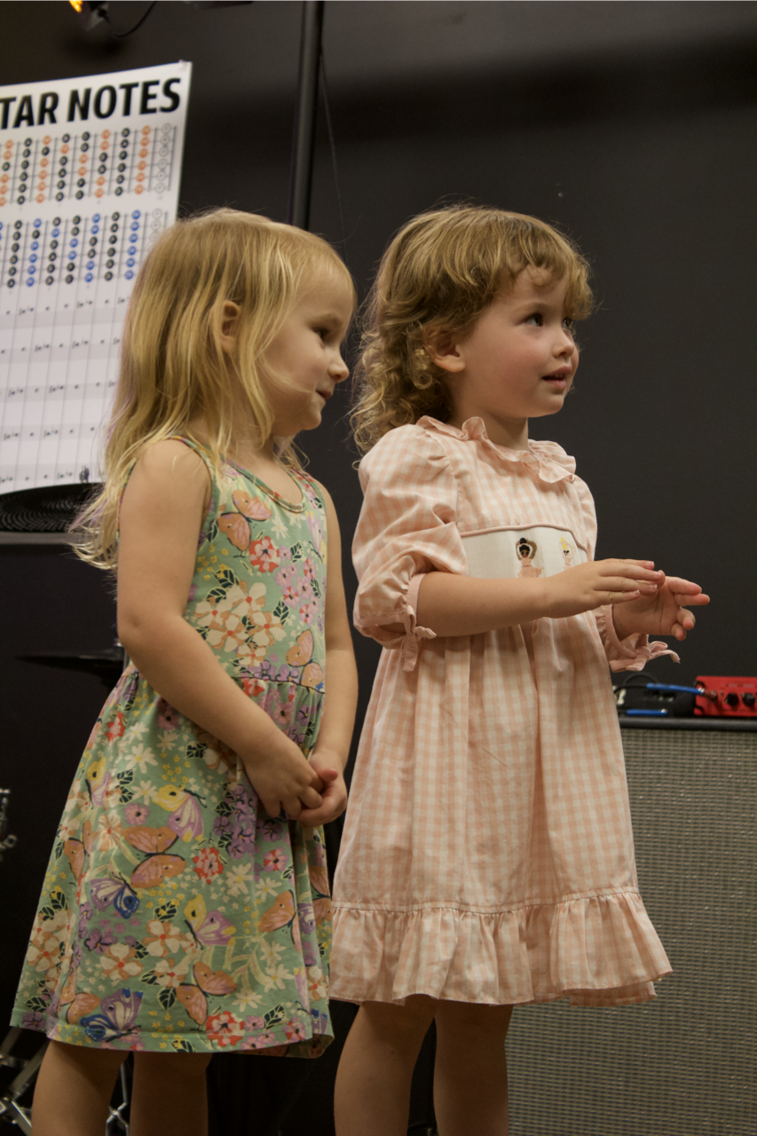 Two young girls standing next to each other, one wearing a floral dress and the other a peach checkered dress, inside a room with a dark wall and a star notes chart in the background.