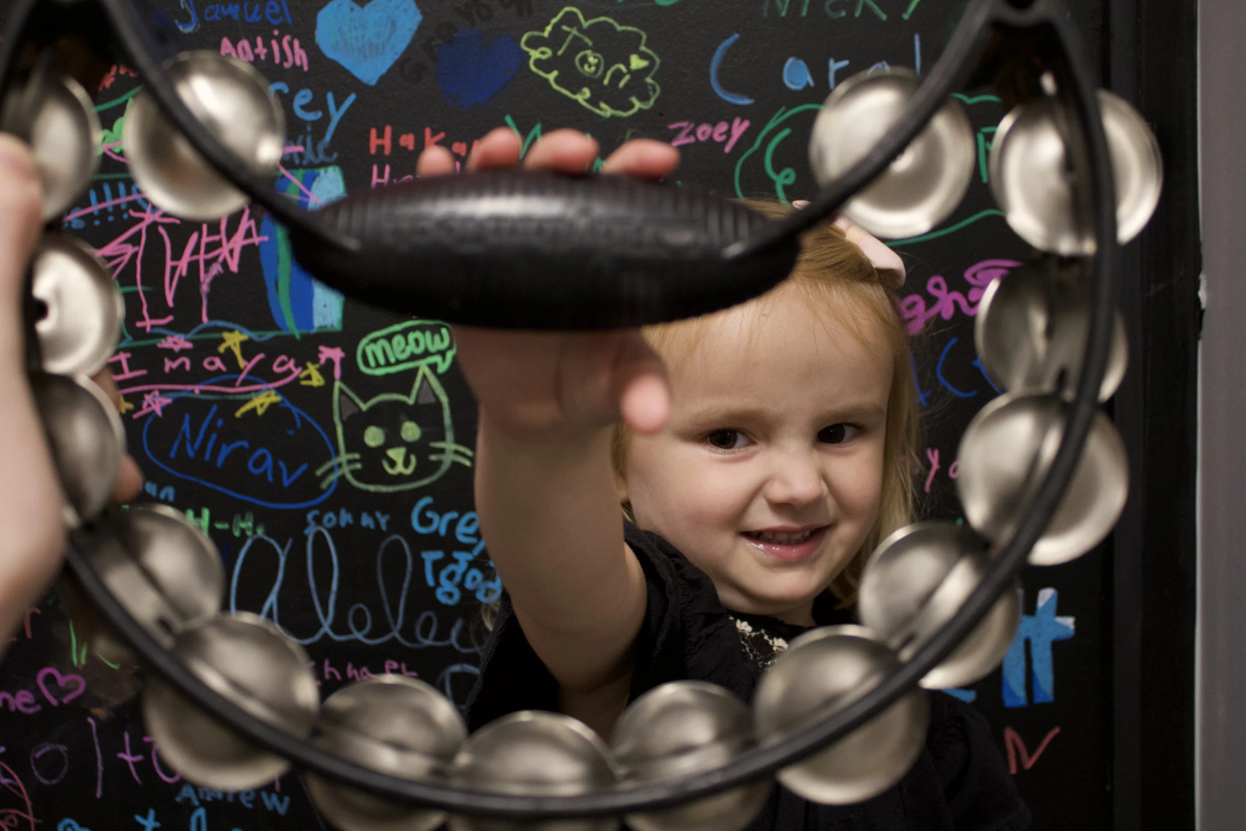 A blonde preschool girl smiles at the camera while holding a Tamborine. She is part of a group music class for ages 3-5 in Cornelius, NC.