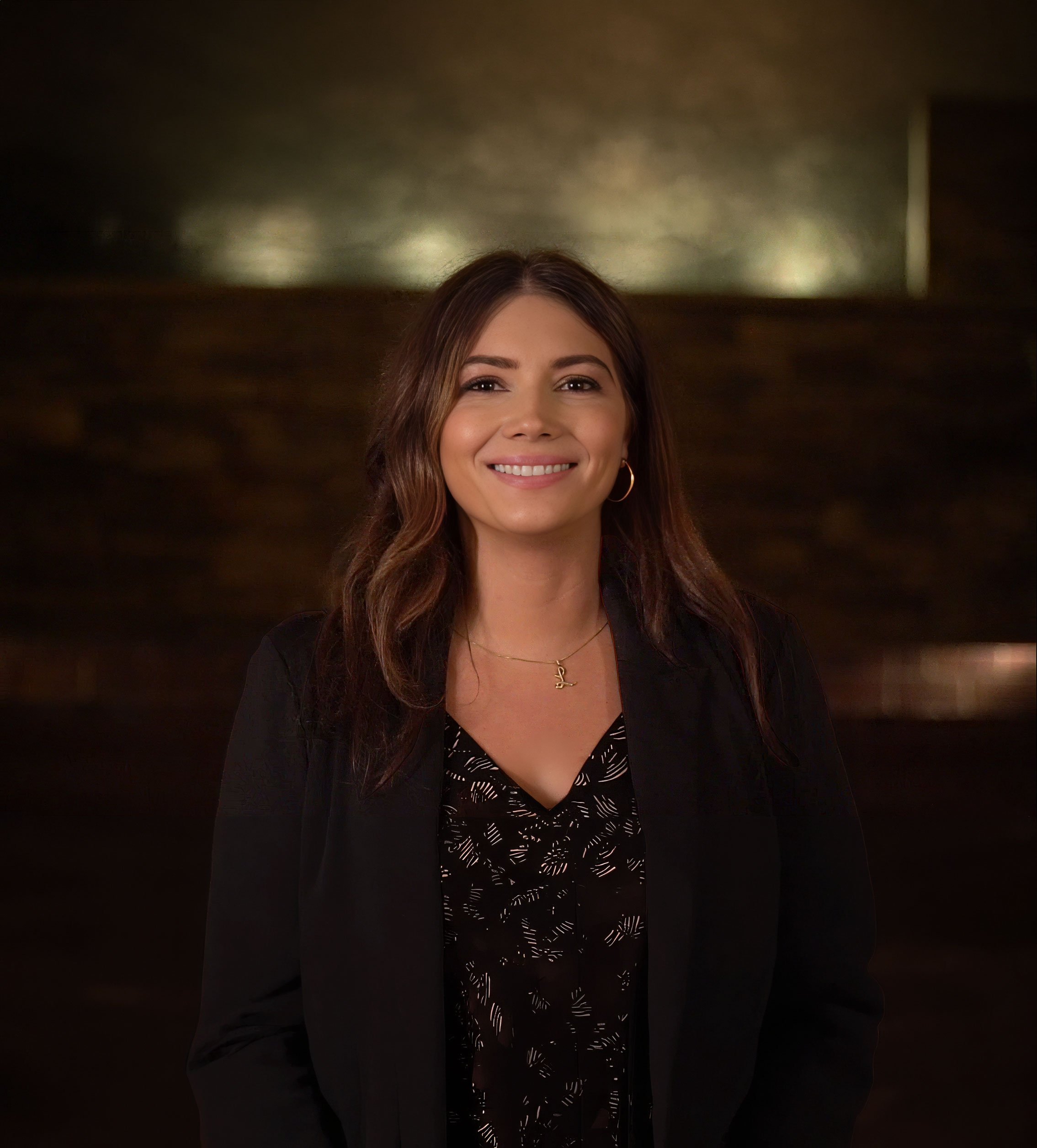 A woman with long dark hair smiling, wearing a black blazer over a patterned blouse, with her hand on her hip, standing against a plain white background.