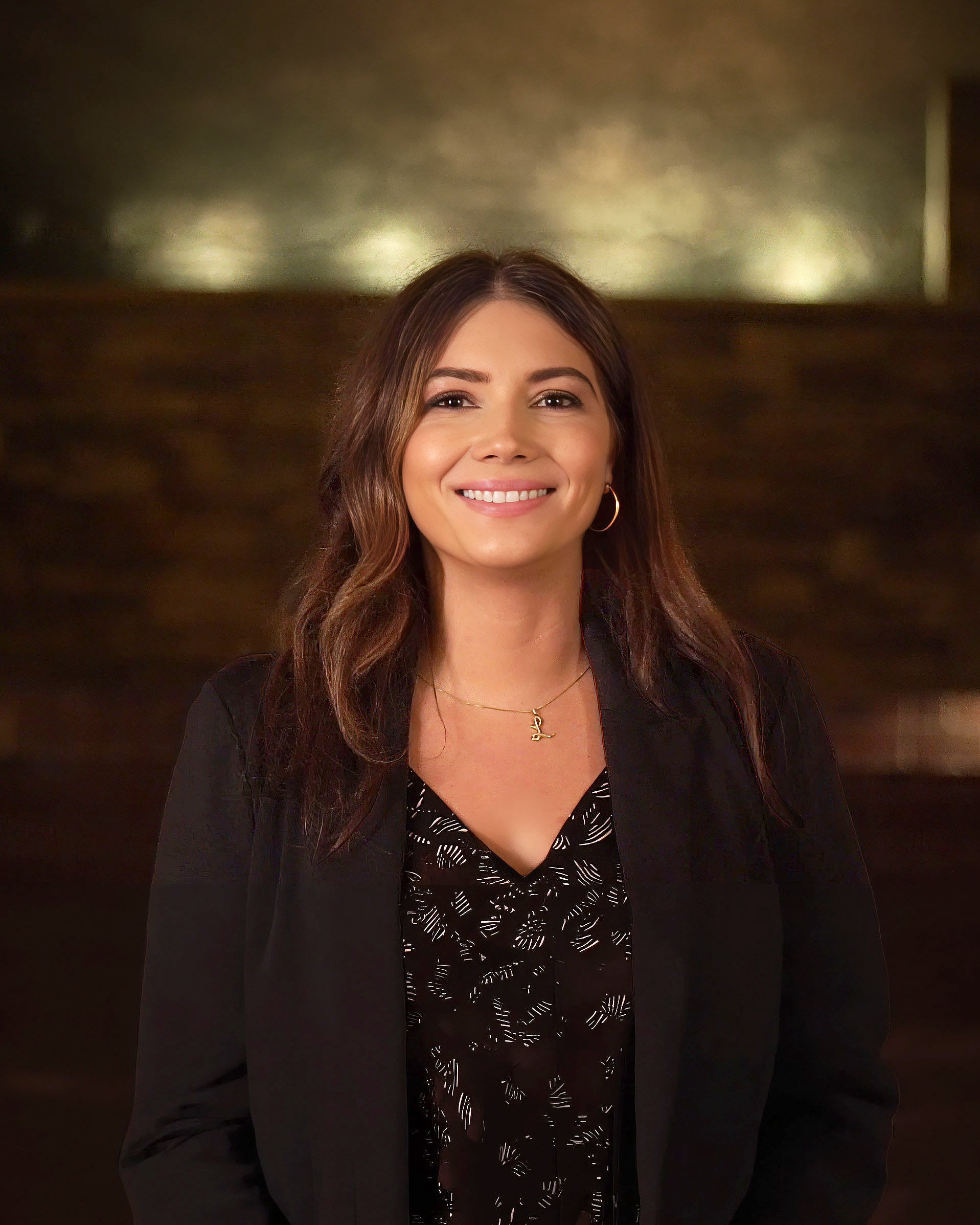 A woman with long dark hair smiling, wearing a black blazer over a patterned blouse, with her hand on her hip, standing against a plain white background.