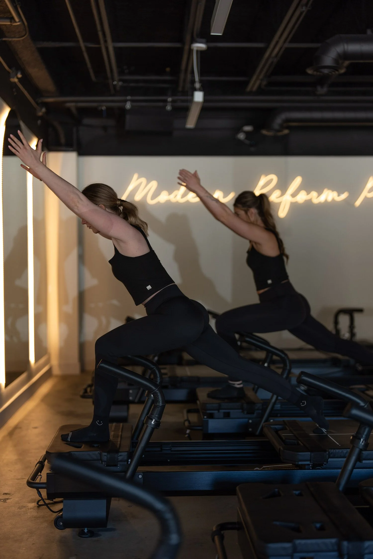 Two women exercising on indoor Pilates reformer machines in a fitness studio with neon sign on the wall.