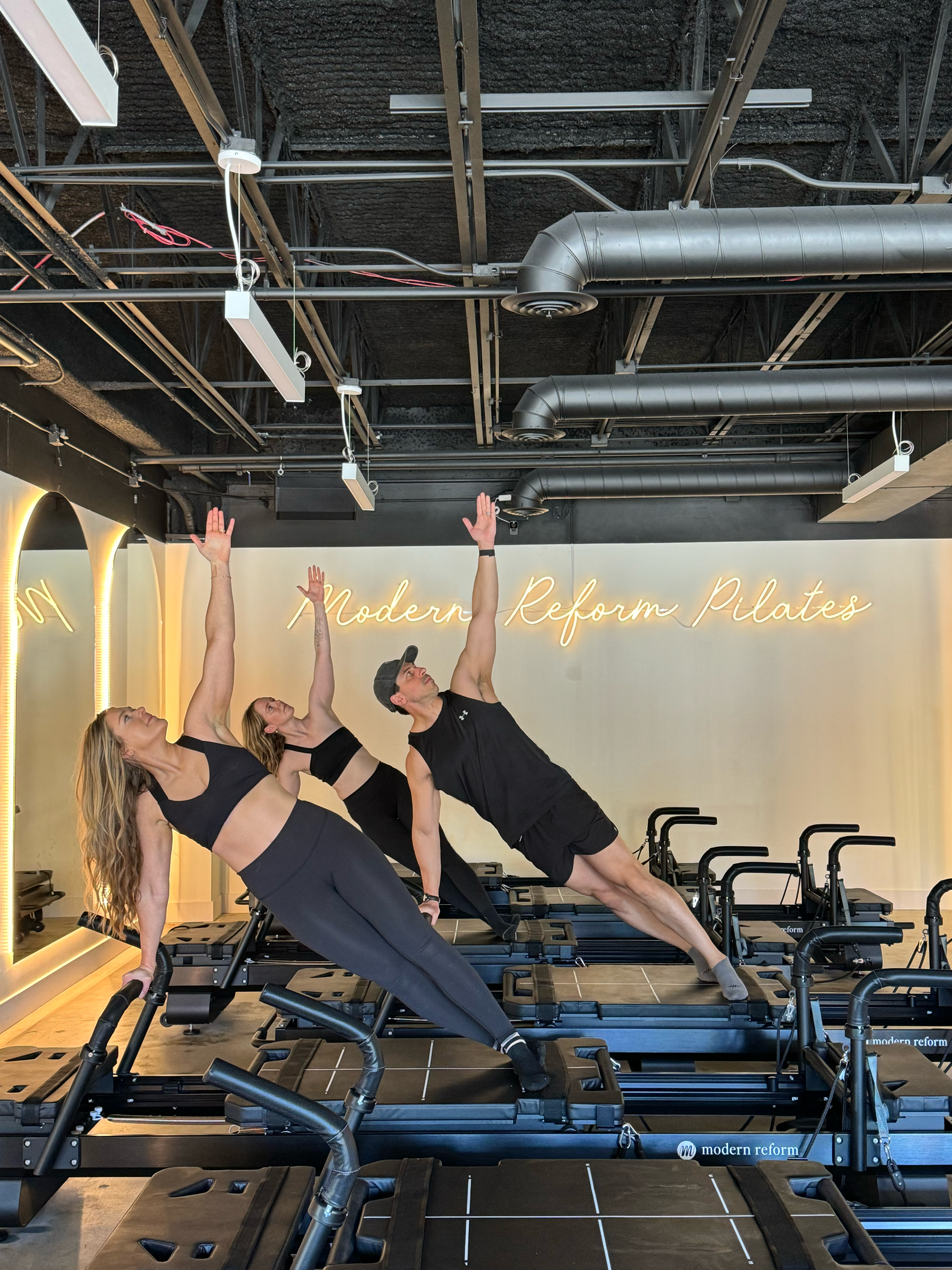 Four people in workout clothes doing side plank exercises on reformer Pilates machines in a modern fitness studio.
