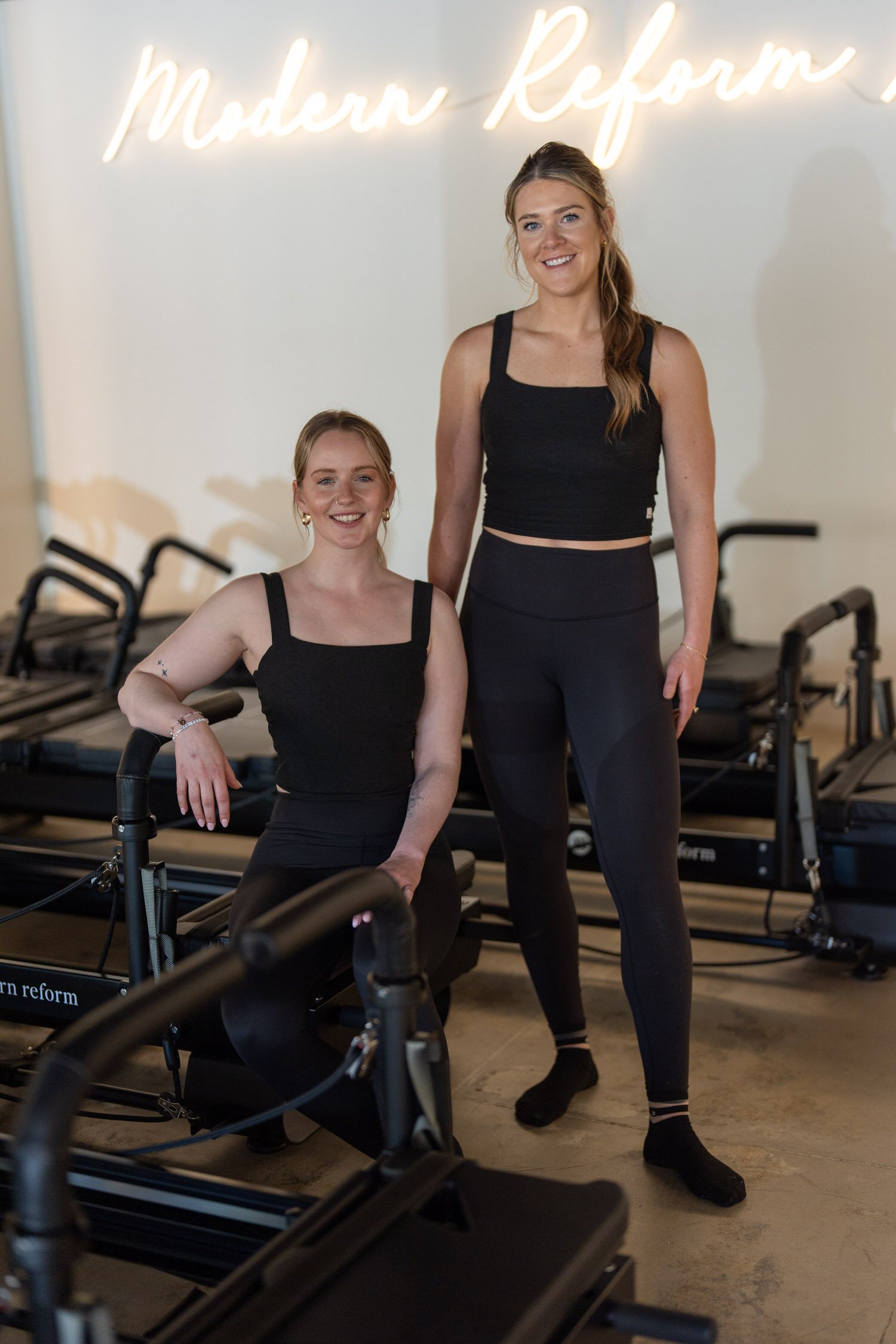 Two women in black workout attire inside a Pilates studio with reformer machines. One woman is standing, and the other is sitting on a reformer machine. A neon sign on the wall reads 'Modern reform'.