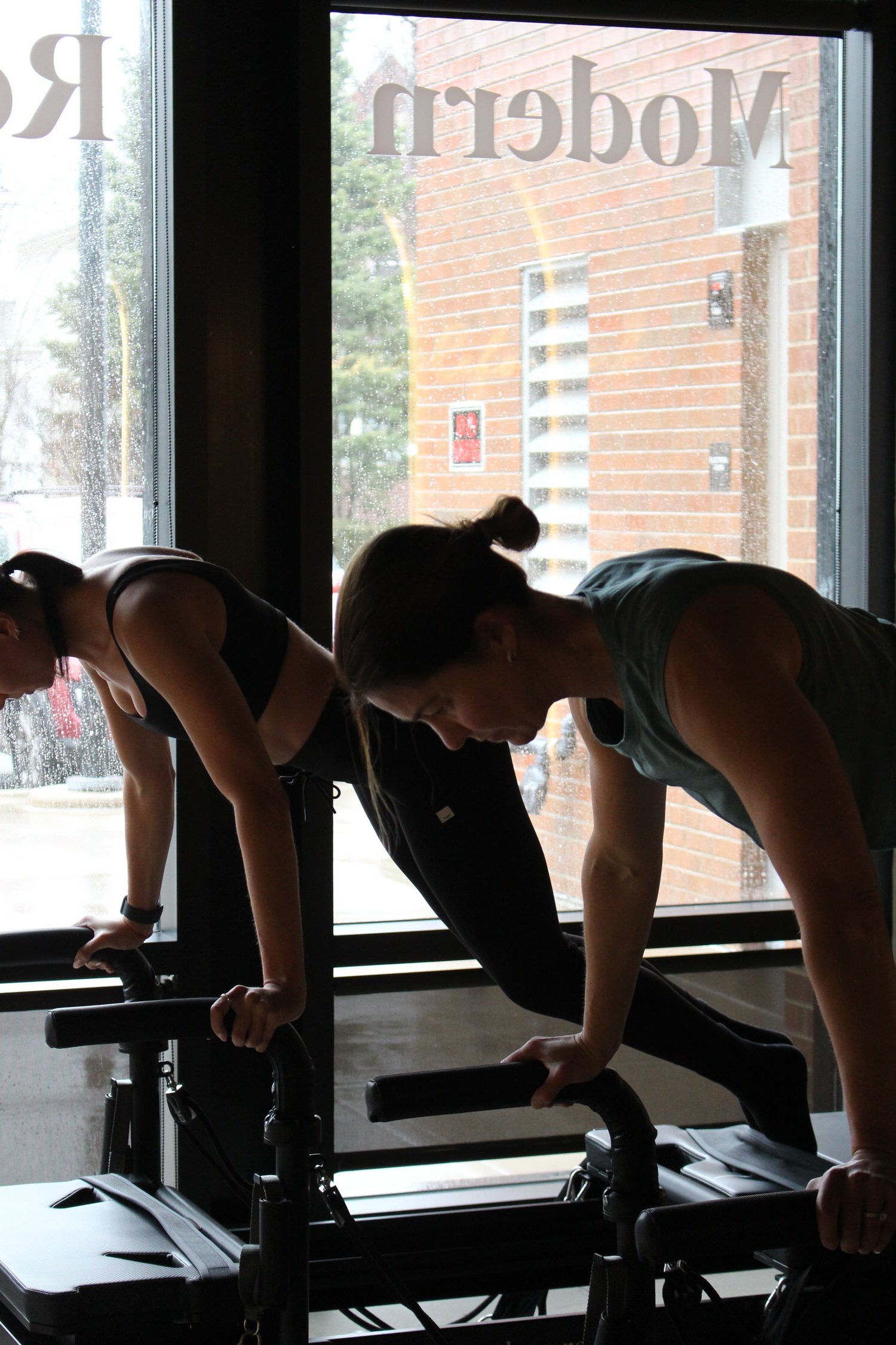 Two women in workout clothes performing planks on stationary exercise bikes inside a gym near large window with rain outside.