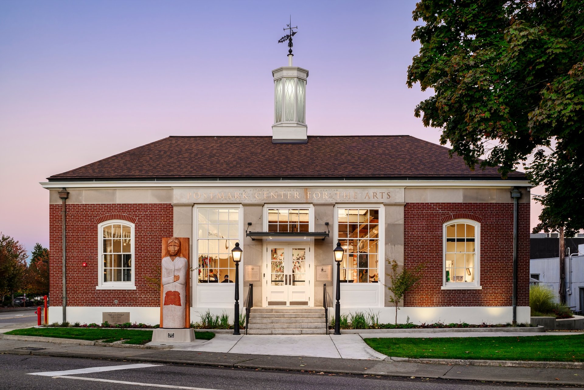 Postmark Center for the Arts building with brick facade and large windows, a sculpture in front, and lit street lamps at dusk.