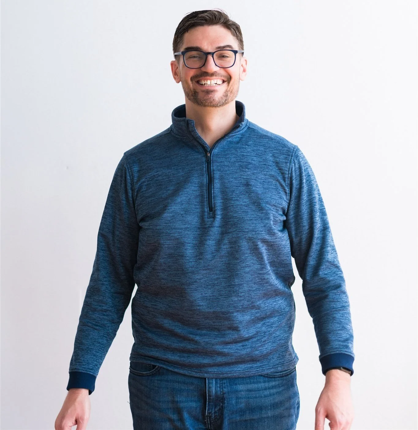 Man with dark hair, black framed glasses, and facial hair smiling with his teeth. He's wearing a dark blue half zip pullover and blue jeans