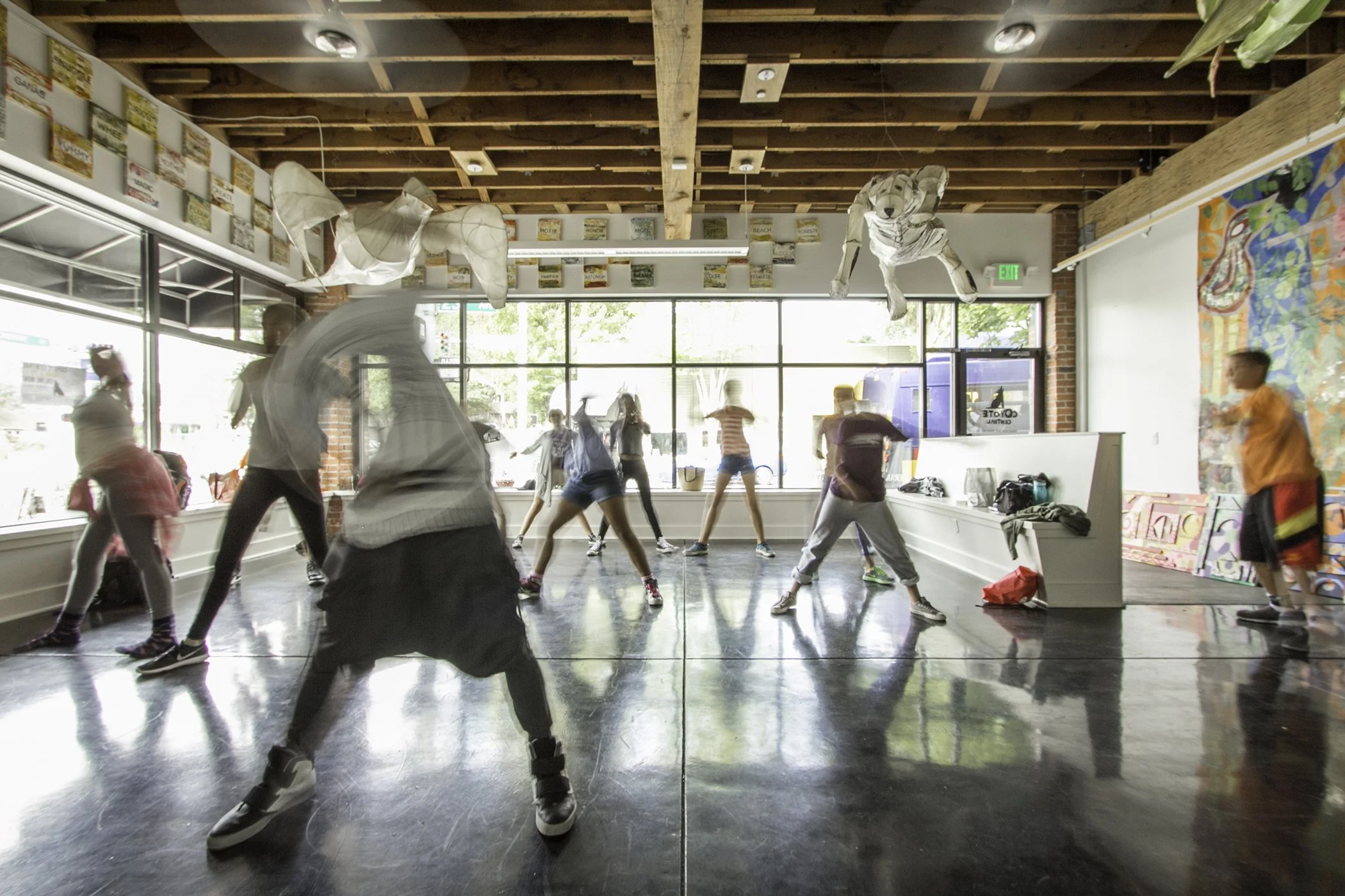 Group of people dancing in a studio with a polished floor, large windows, and hanging decorations.