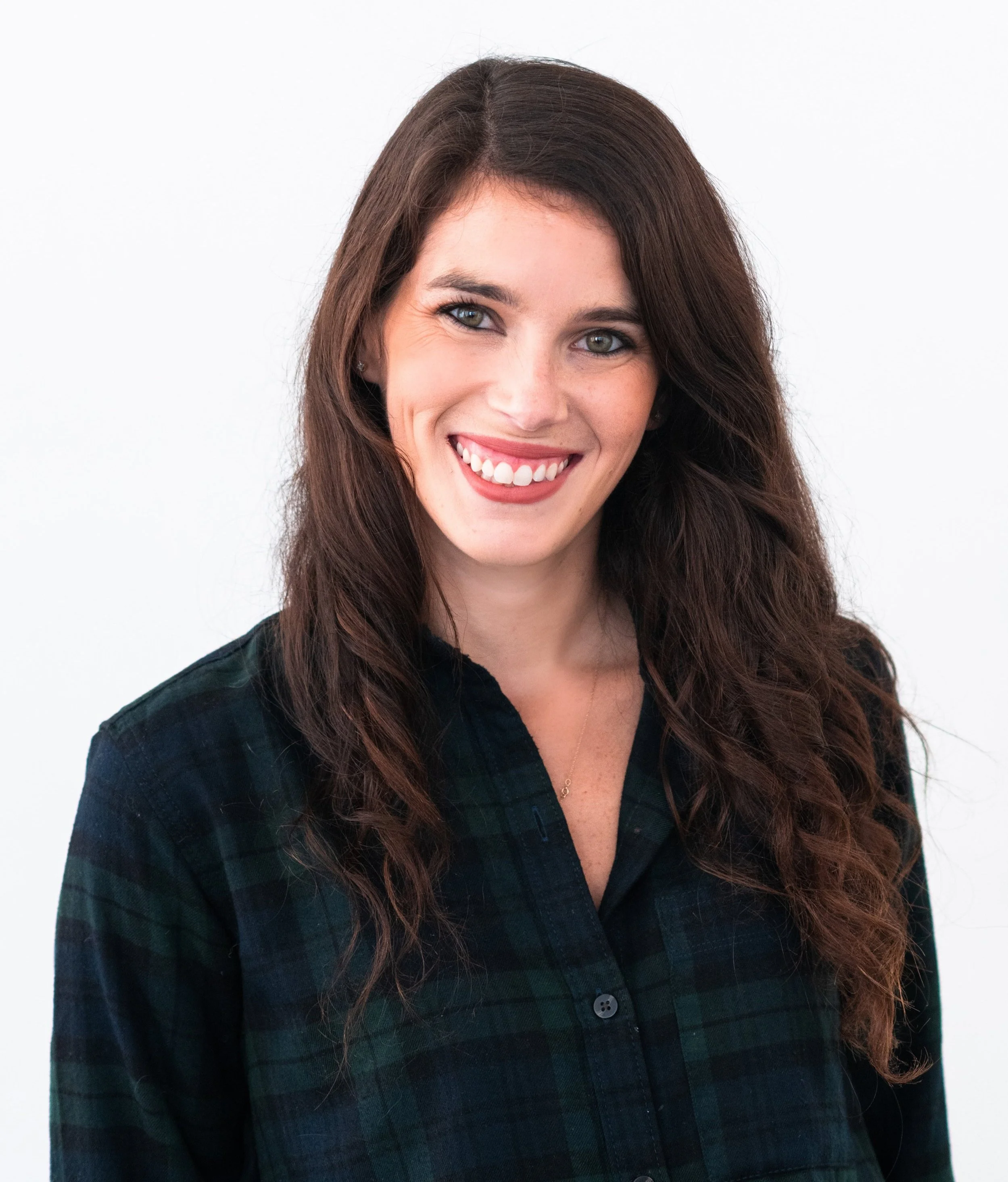 Headshot of a woman with long dark hair and smiling with her teeth.  She's wearing a dark green and navy plaid button-up shirt.