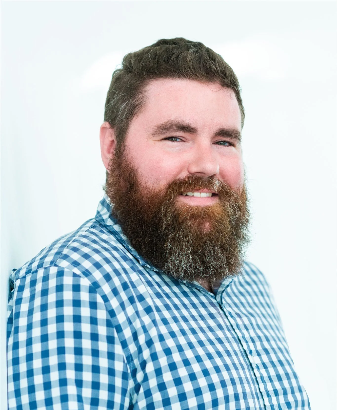 Headshot of a man with brown hair and large brown beard with some grey hairs. He is smiling with his teeth and is wearing a white and blue check plaid shirt.