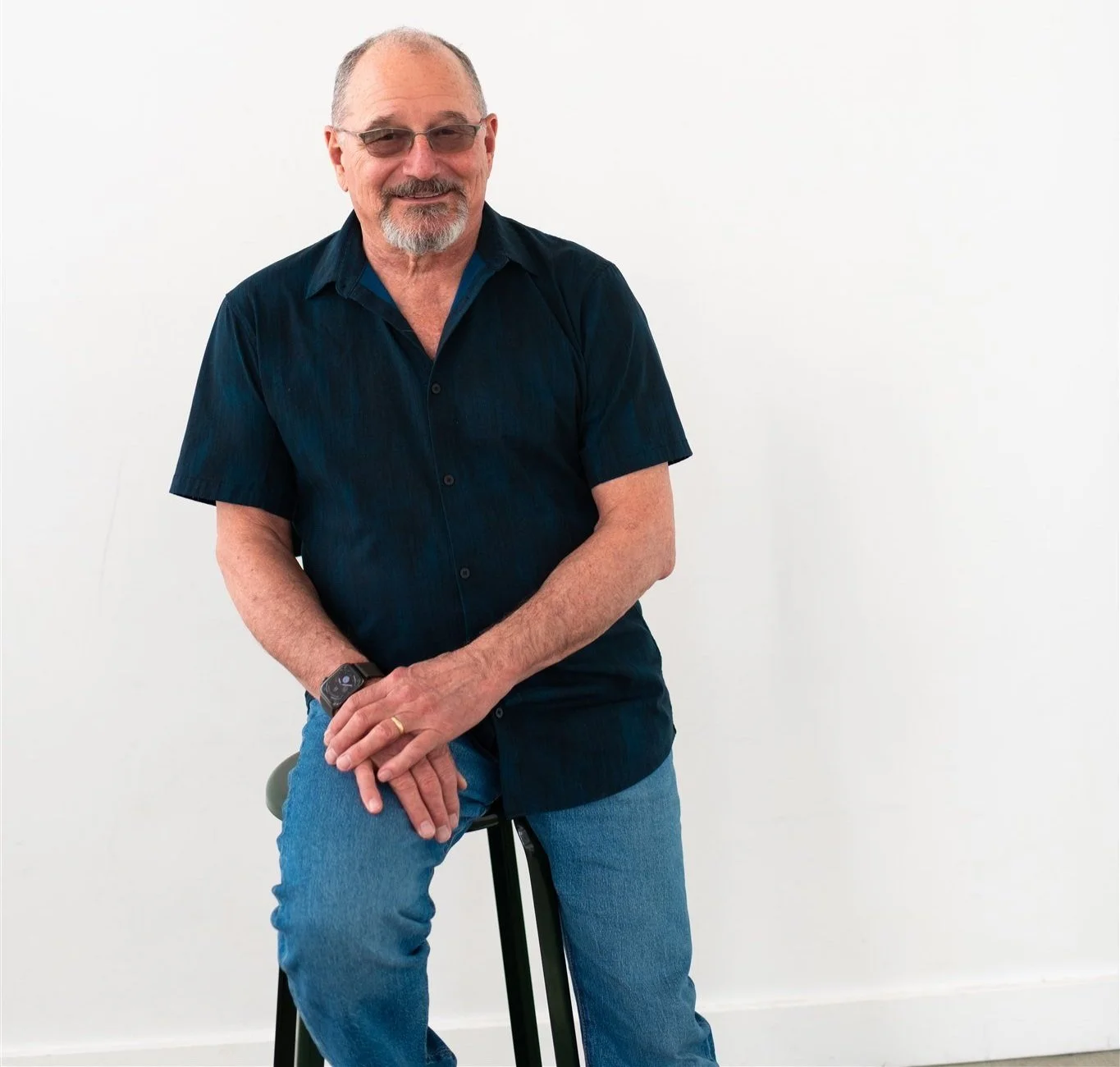 Man sitting on a stool and leaning to one side. He's wearing transition lens glasses, a navy short-sleeve button-up  shirt and blue jeans.