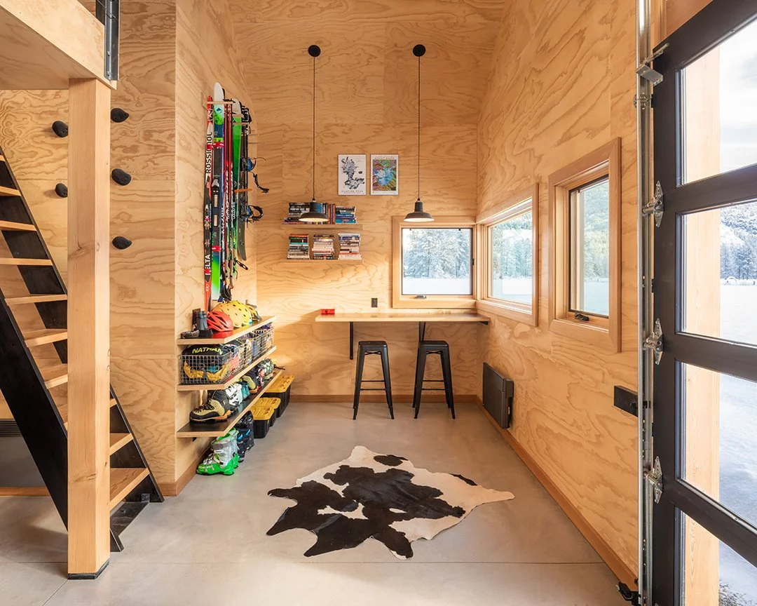 Inside of a small gear shed with wood paneled walls. There is ski gear hanging on a wall, a built-in bar with two stools that look out through the windows to the snowy outdoors, and a stair ladder with climbing holds along the wall.
