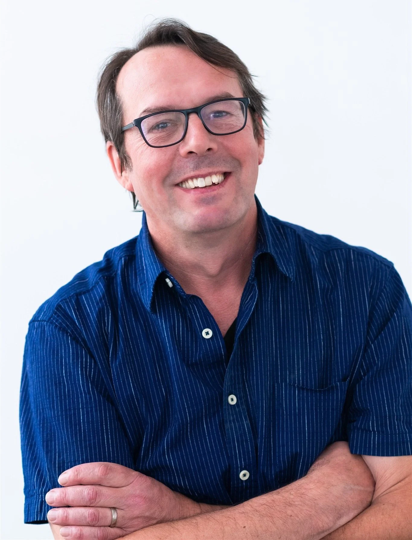 Man with dark hair and black framed glasses smiling with his teeth. He's wearing a dark blue and white pinstripe button-down shirt with his arms crossed.