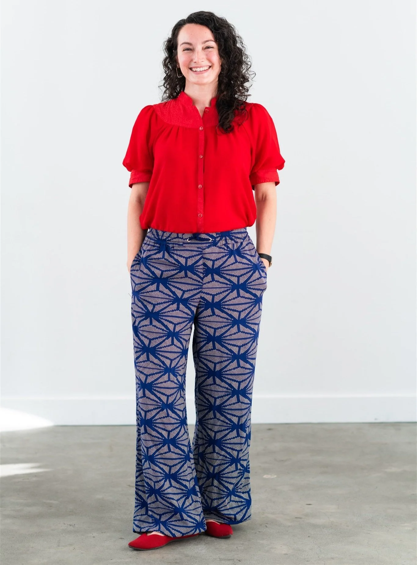 Woman with dark hair smiling with her teeth and hands in her pockets. She's wearing a red short sleeved blouse and wide-leg pants with a blue pattern.