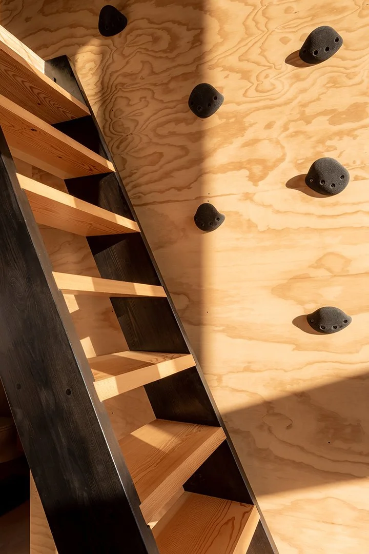 Close-up detail of wooden ladder stairs and the wood panel wall it's against. There are climbing holds attached to the wall.