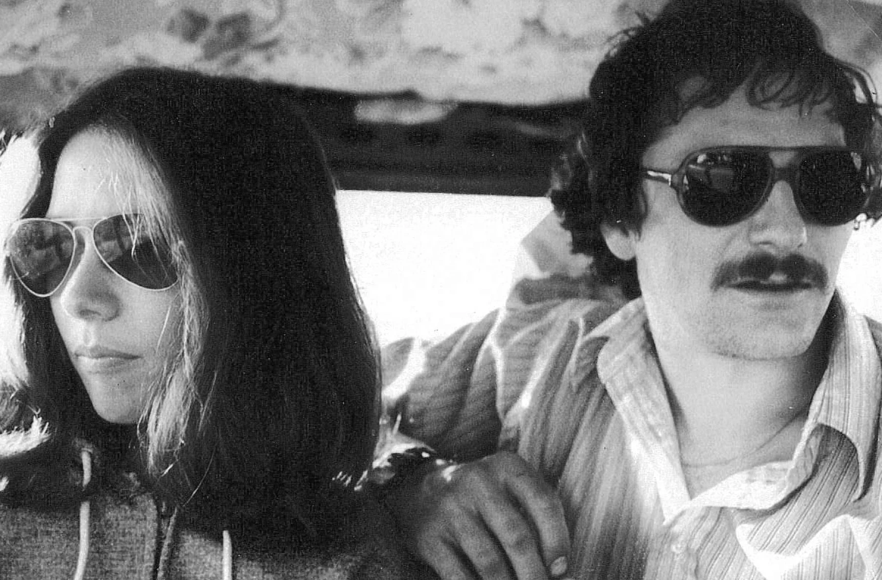 Black and white photo of a man and woman wearing sunglasses, with the man having a mustache.