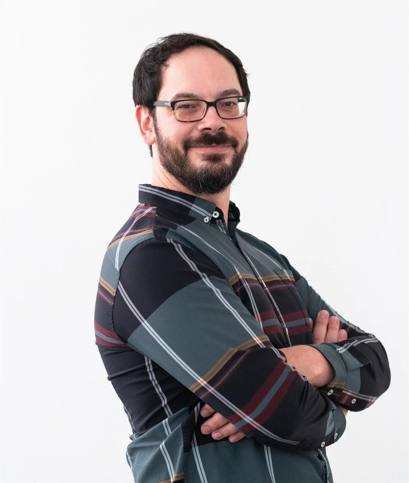 Headshot of a man with dark hair and beard and arms crossed. He's wearing black frame eyeglasses and a blue grey plaid shirt.