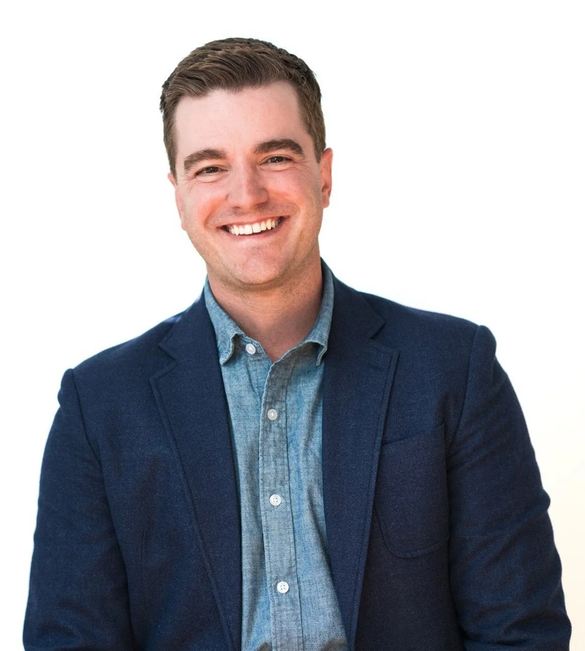 Man with short brown hair smiling with his teeth and wearing a chambray button-up shirt under a navy blazer.