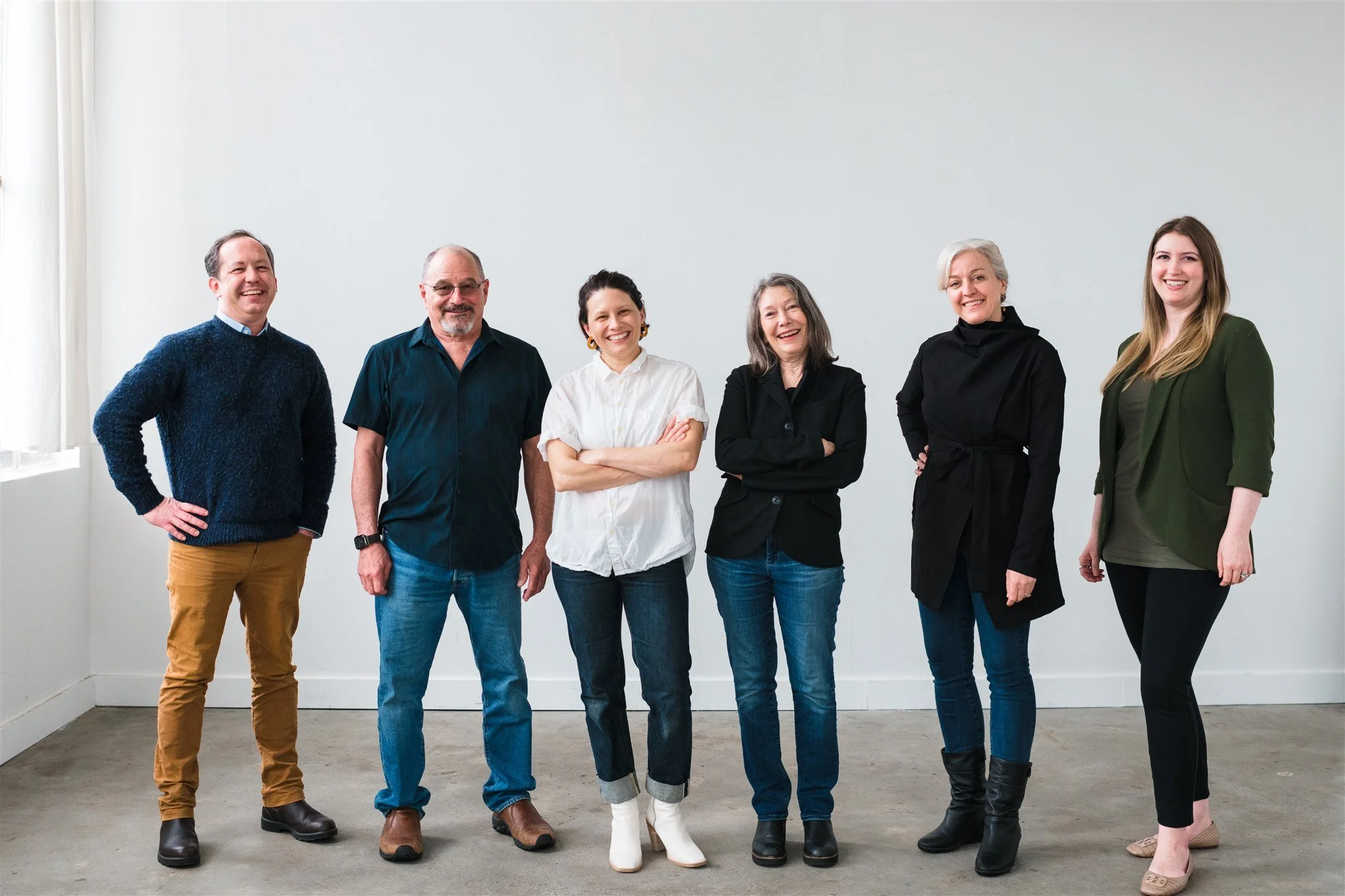 Group of six people standing against a white wall, casually dressed, smiling.