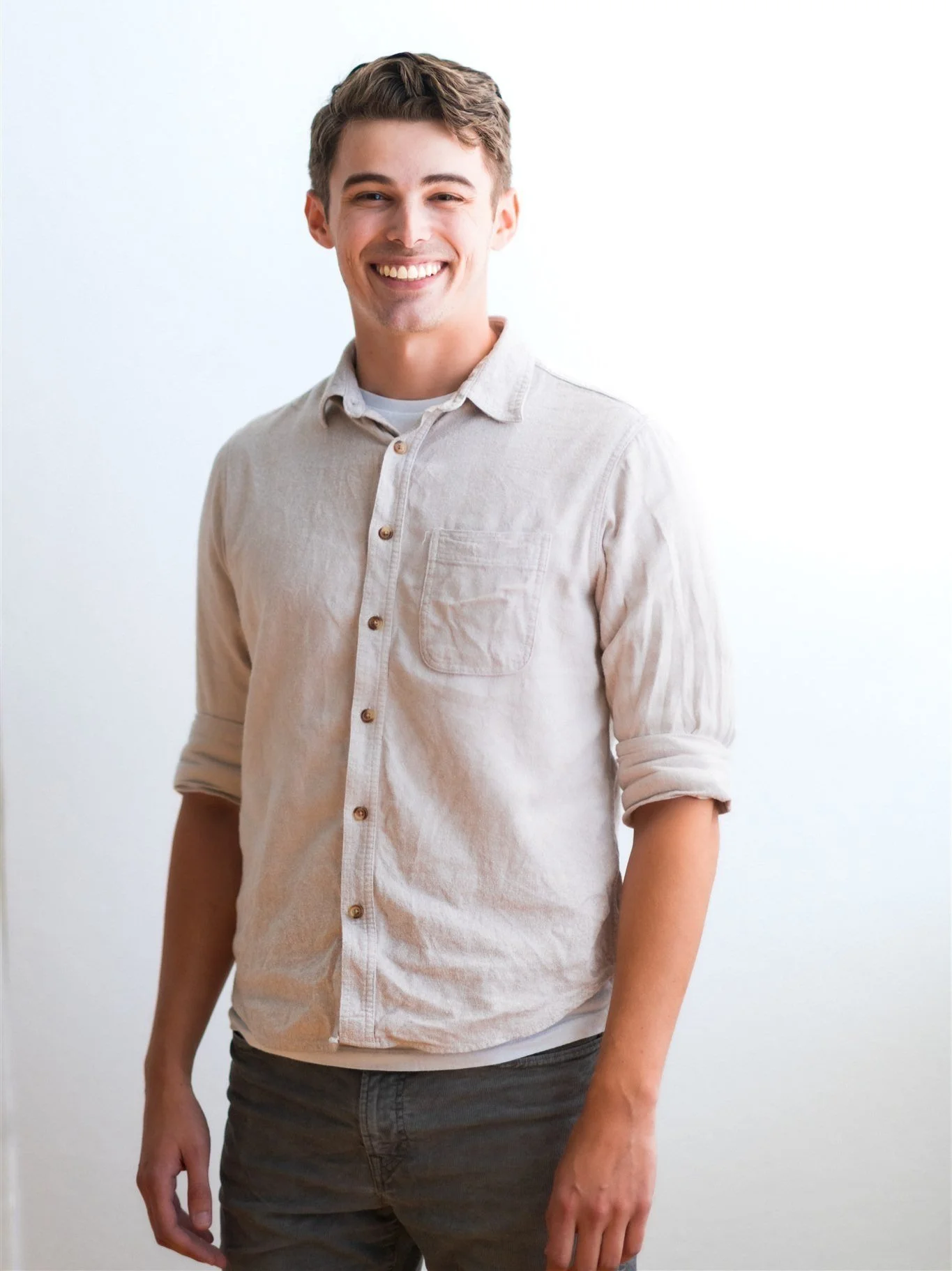 Smiling person wearing a beige button-up shirt with rolled sleeves against a white background.
