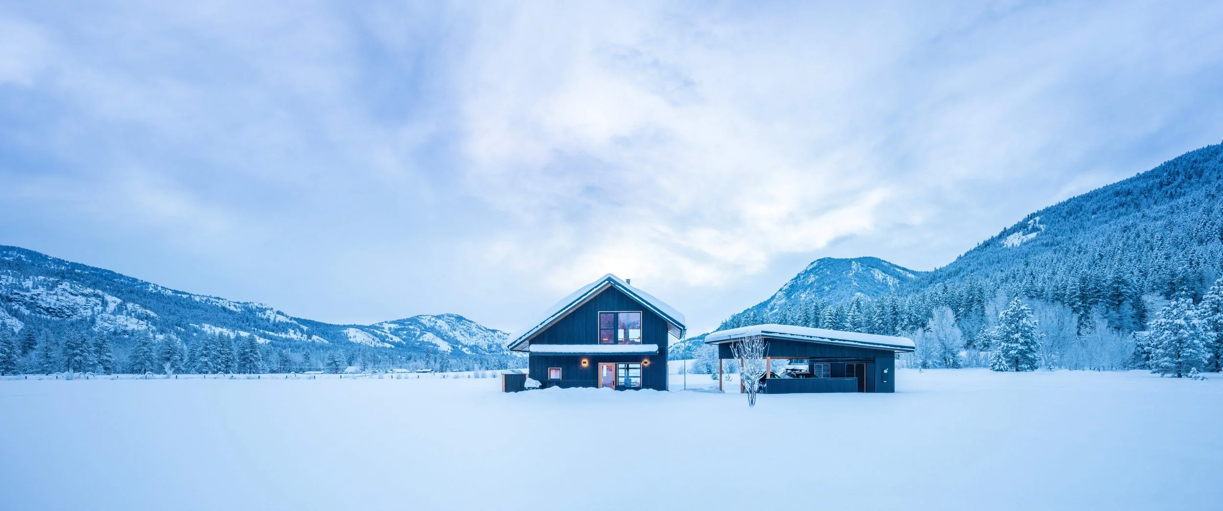 Small house and a detached open garage in the middle of a snow covered valley. Behind the buildings is a tree scaped mountain range.