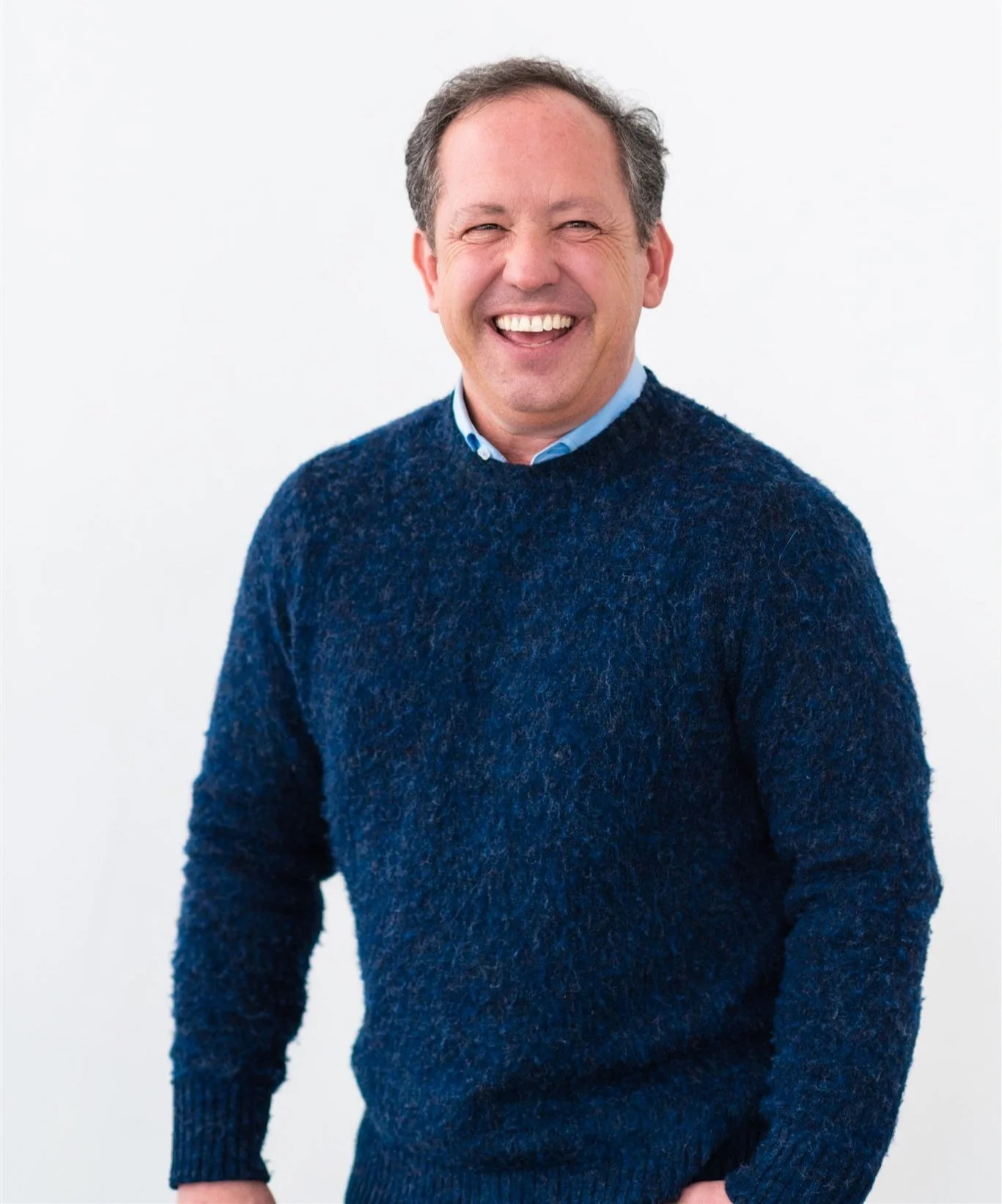 Man with greying hair smiling with his teeth and wearing a dark blue sweater with a light blue collard shirt underneath.