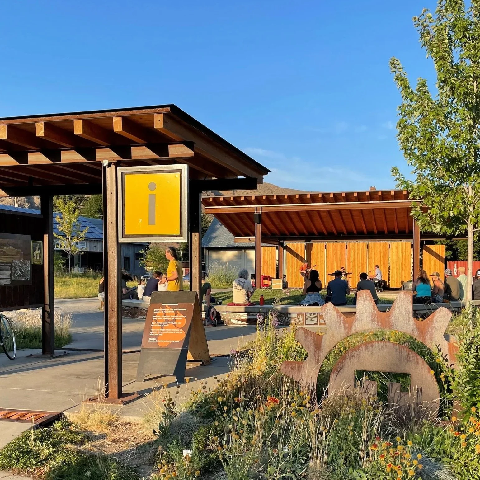 Outdoor seating area with wooden shelters, information sign, bicycle, and people gathered at a table. Surrounded by greenery and flowers, with a metal art installation resembling a gear.