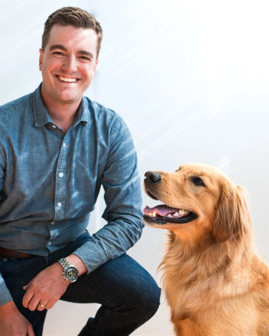 Smiling man in blue shirt sitting beside a golden retriever against a light background.