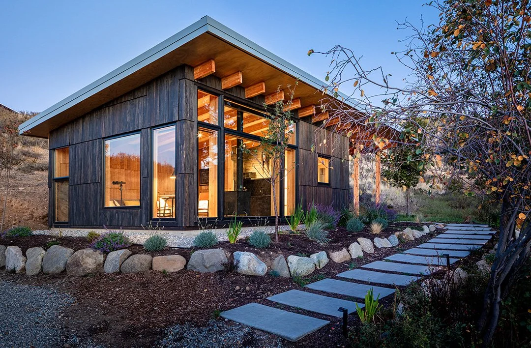 Modern cabin with glass windows, dark wood siding, landscaped garden, stone pathway, and natural surroundings.