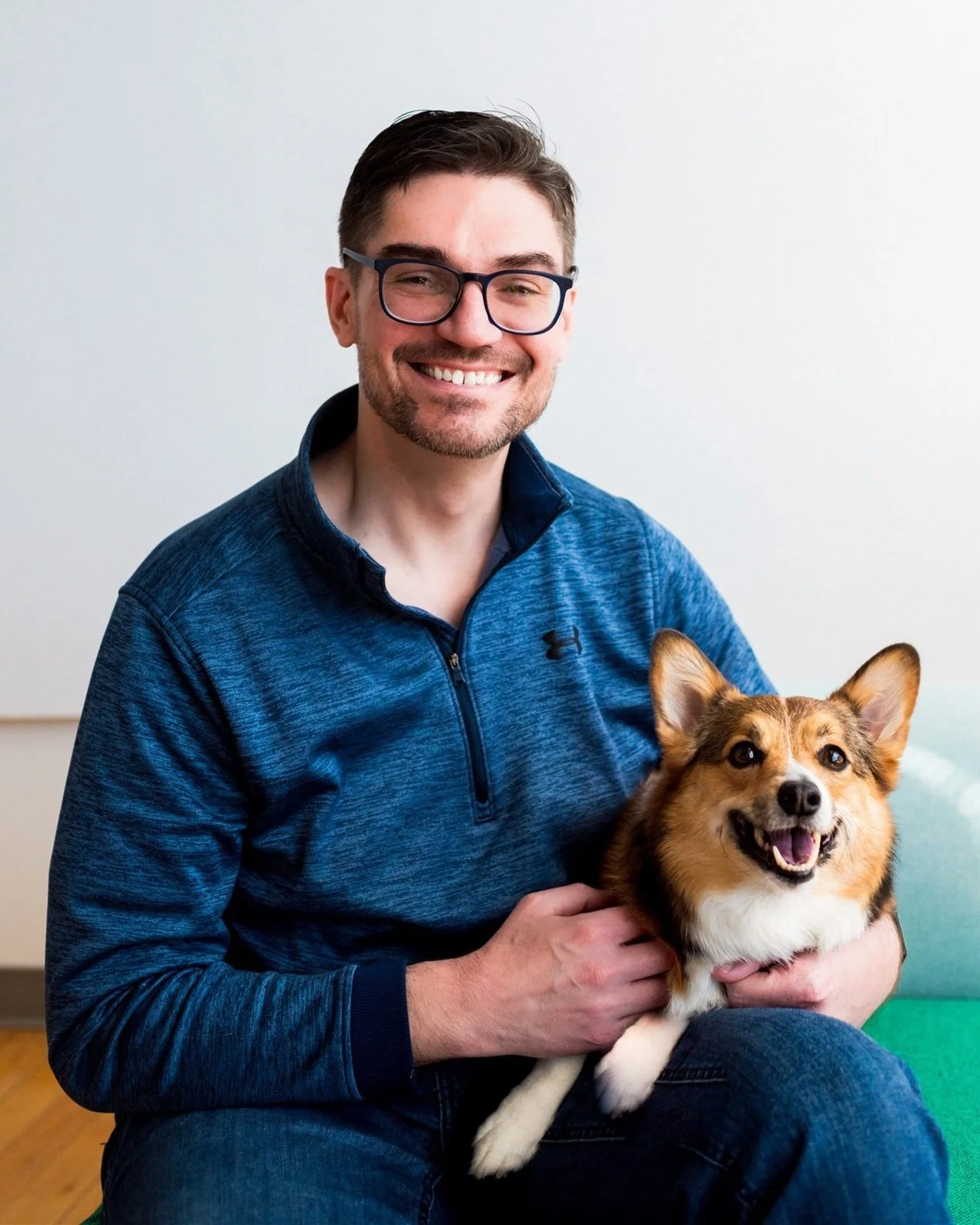 Man with glasses in blue sweater smiling, holding a Pembroke Welsh Corgi dog on his lap.