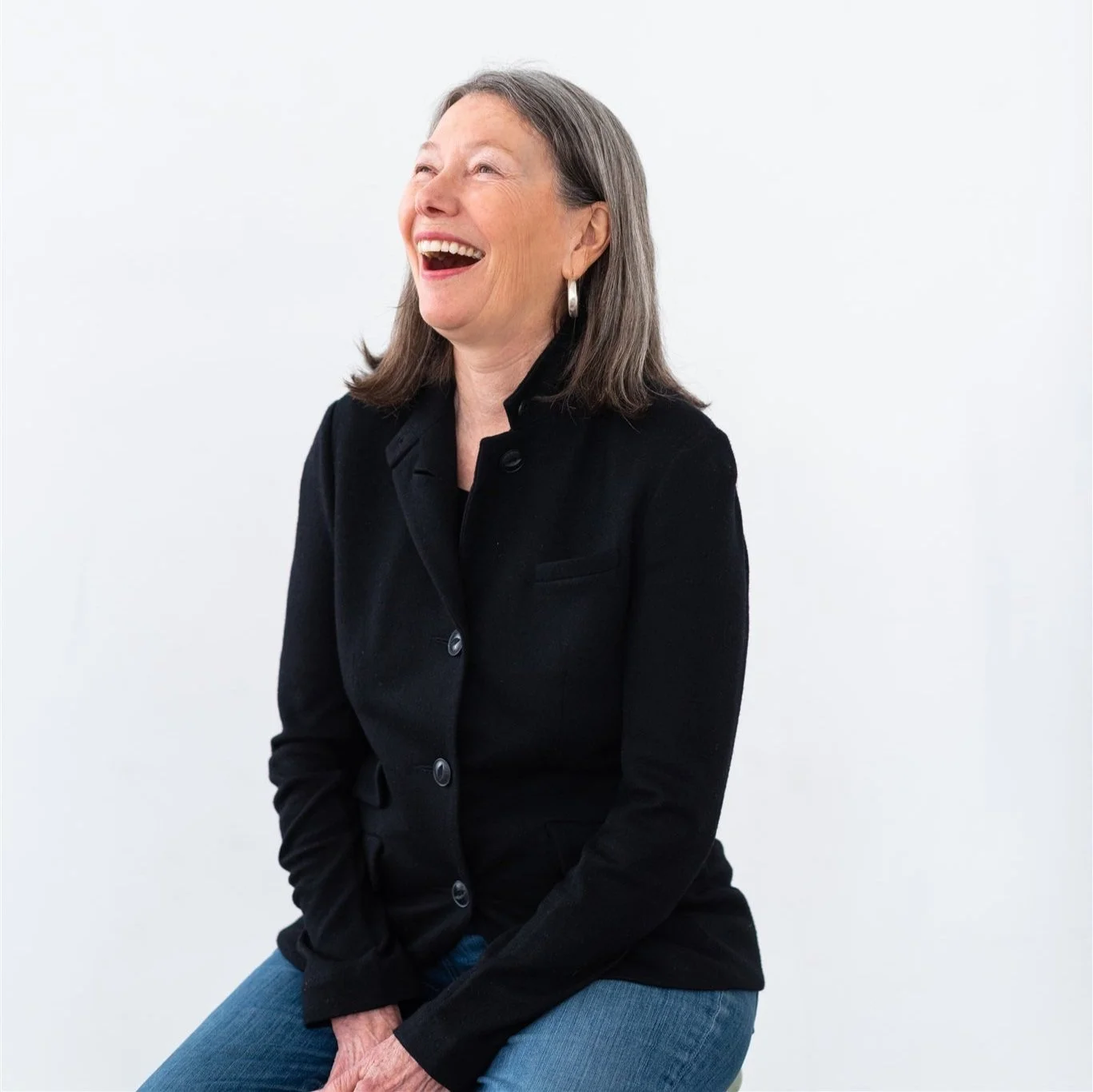 Woman with shoulder-length gray hair sitting on a stool with a huge, open, smile. She is wearing a black button-up jacket and blue jeans.