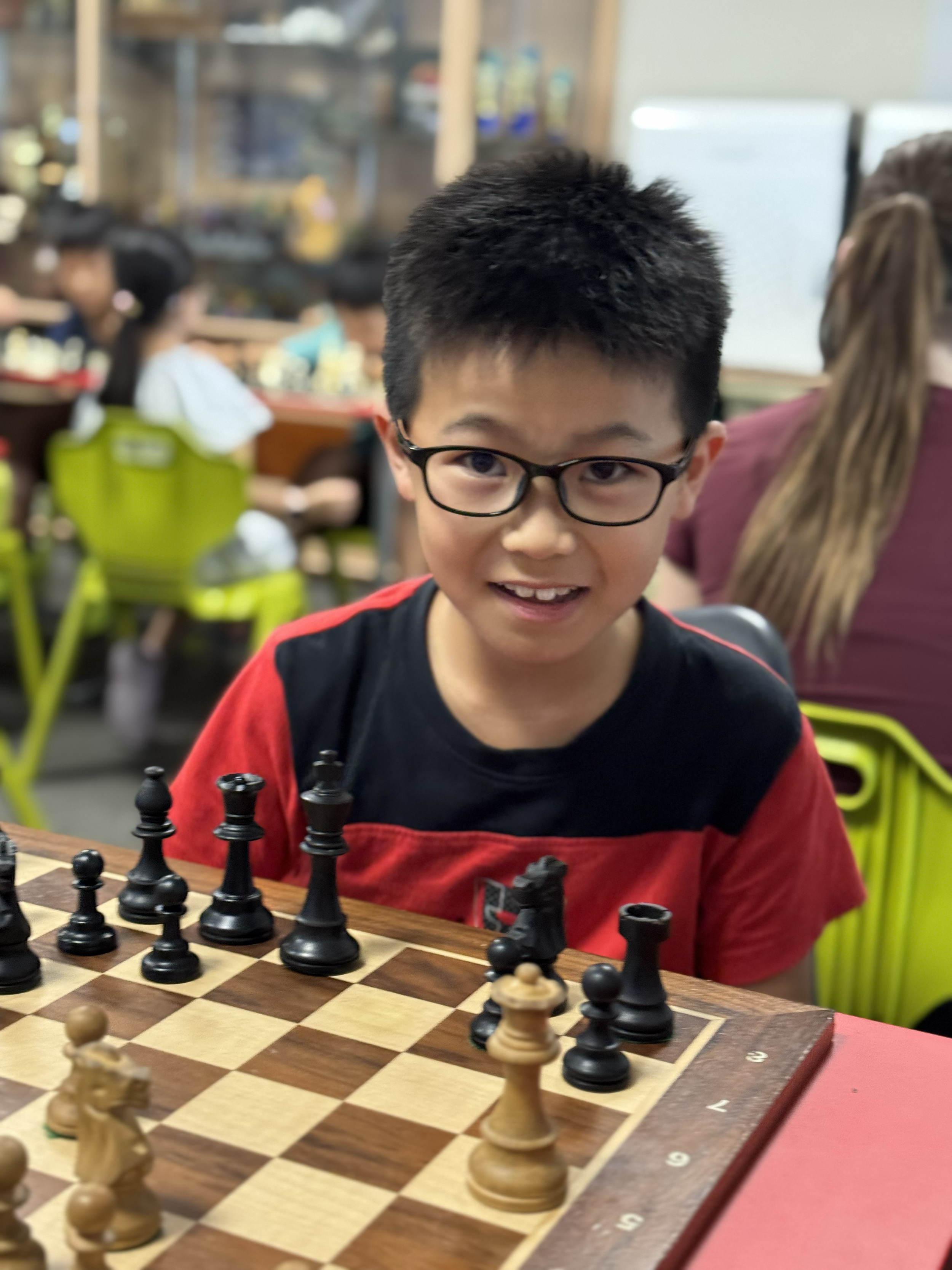 Young boy with glasses smiling, playing chess in a busy indoor setting with other people in the background.