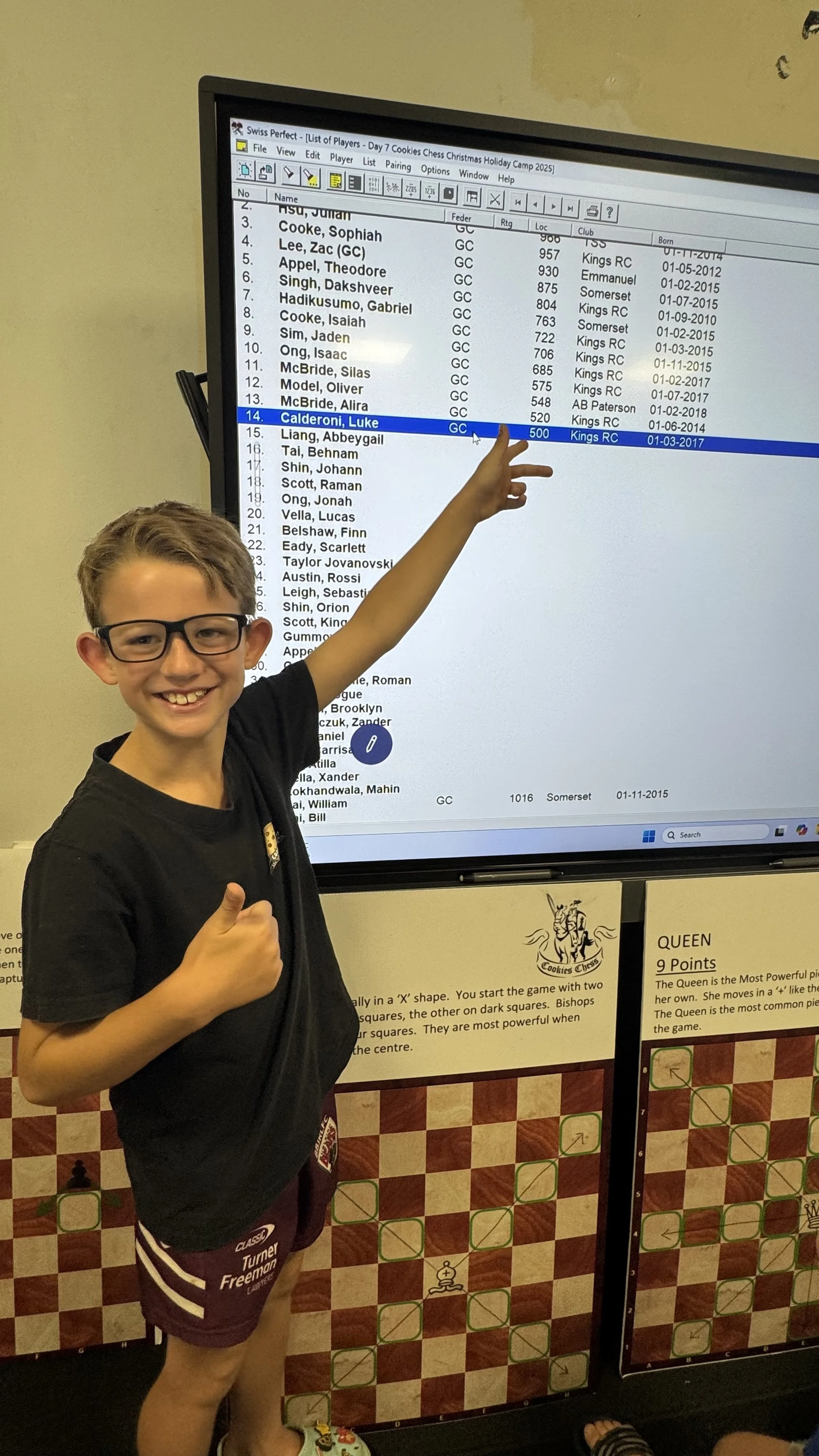 A young boy wearing glasses and a black T-shirt smiling and pointing at a digital chess player list on a screen. The list shows names, team, rating, and birthdate details. Below, there are chessboard displays with instructions for chess game rules.