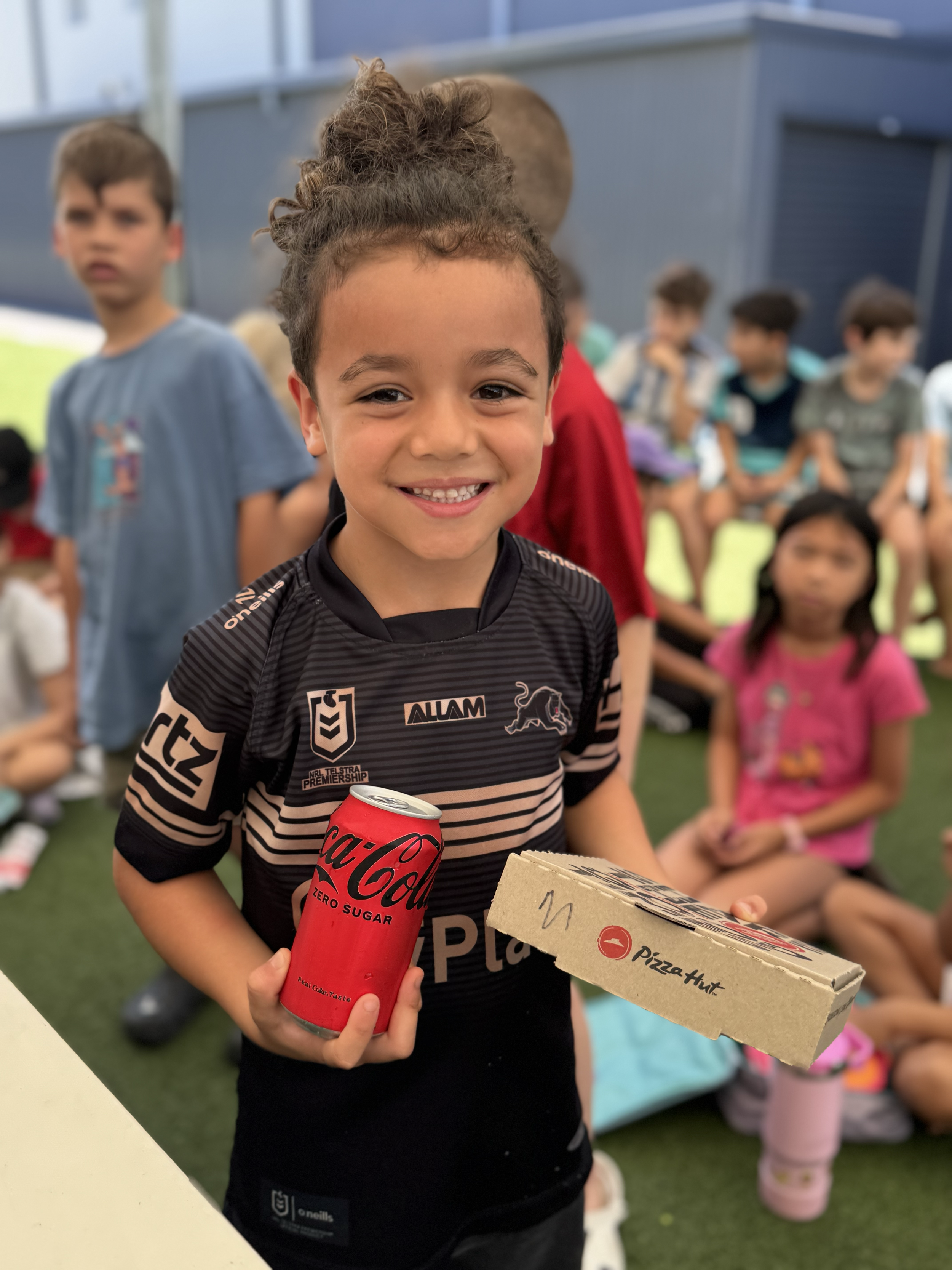 A smiling young boy holding a 12-pack of Coca-Cola Zero Sugar and a pizza box at a gathering of children outdoors.