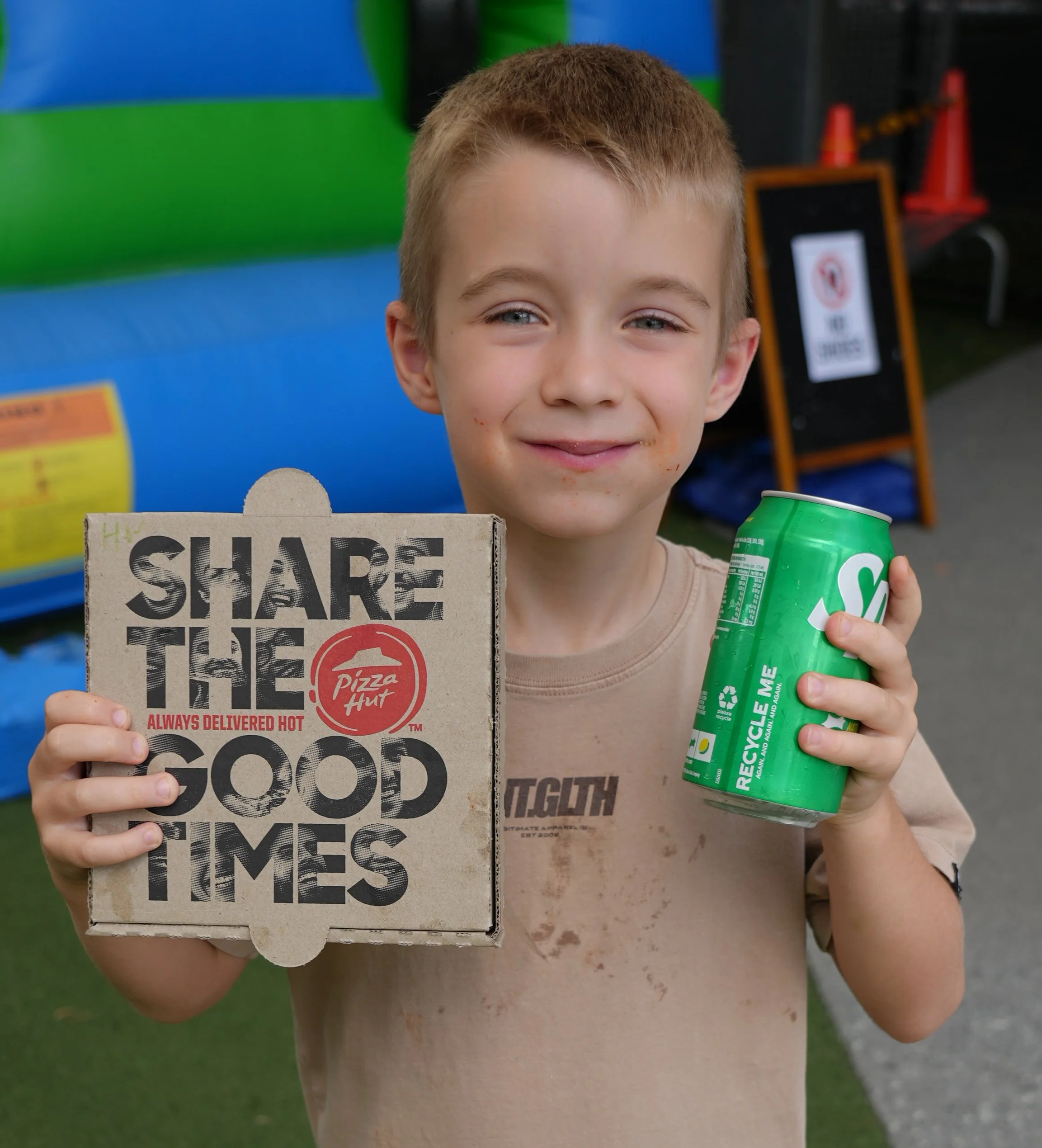 A young boy with a slight smile holding a pizza box with the message 'Share the Good Times' and a green can of Sprite, standing outdoors near a bounce house and orange cones.