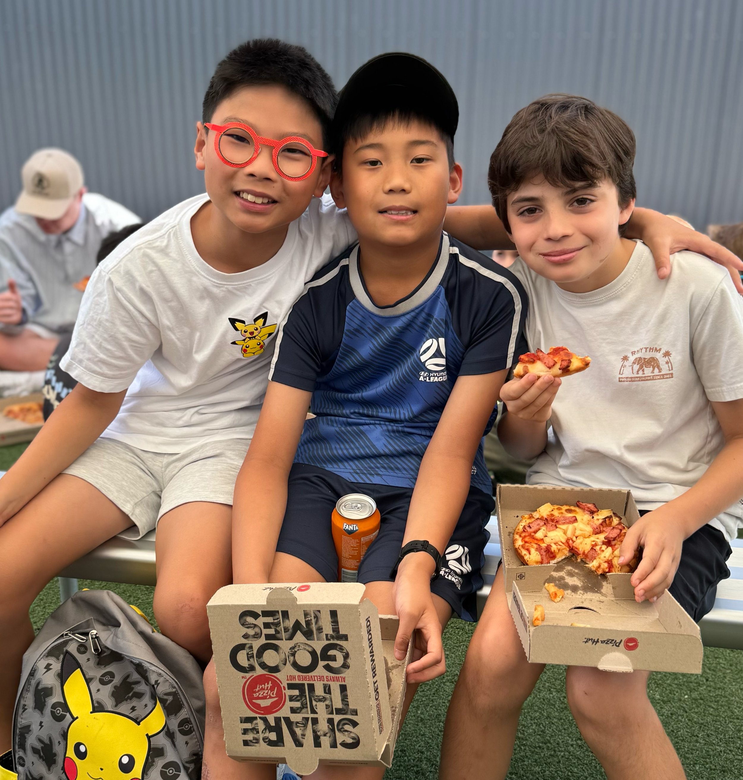 Three boys sitting together, holding pizza slices and pizza boxes, with a backpack featuring Pikachu in front of them.