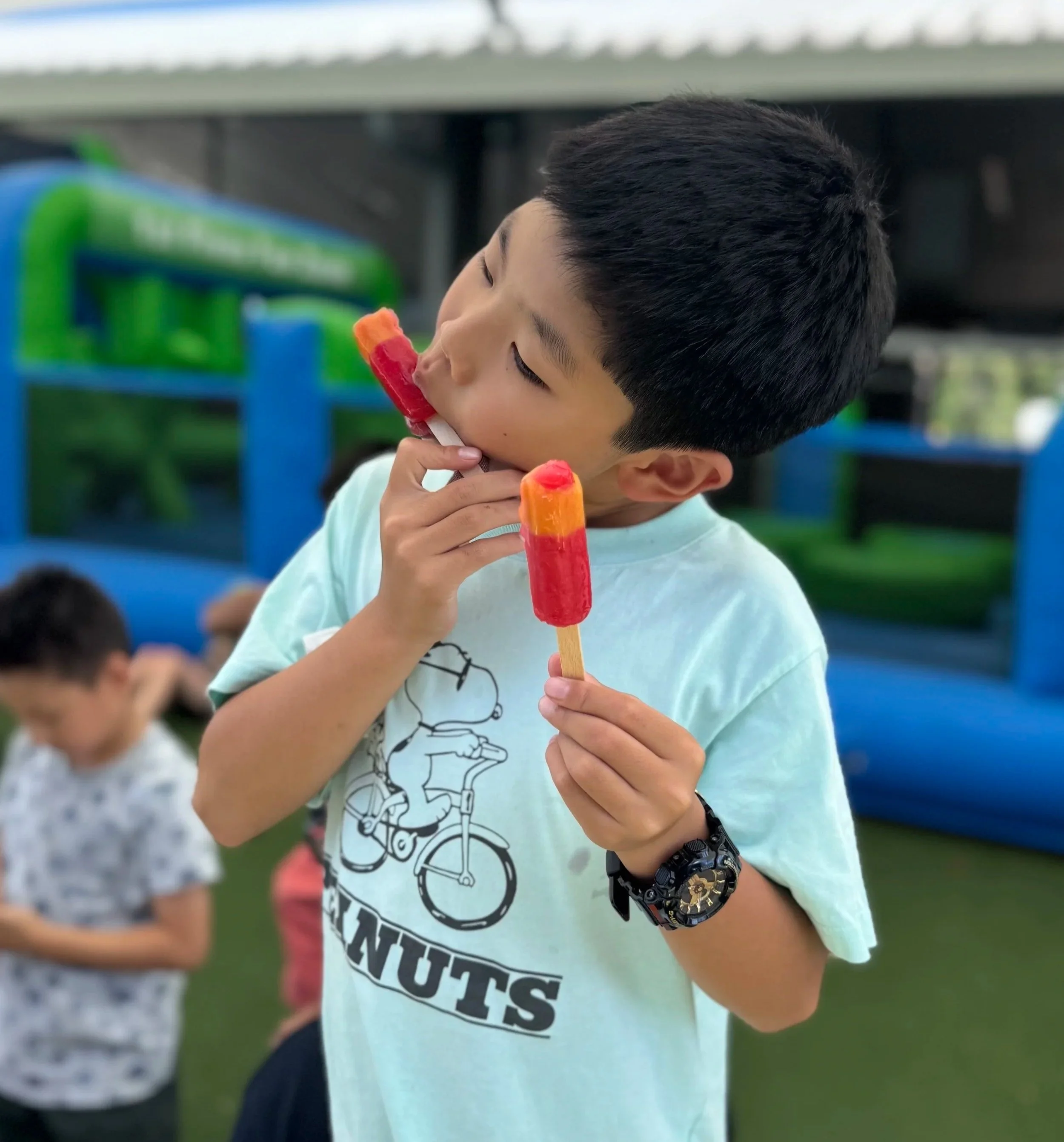 A boy with short black hair and a watch on his wrist, wearing a light blue t-shirt with a cartoon character riding a bicycle and the text 'NUTS', is holding three ice pops and licking one.