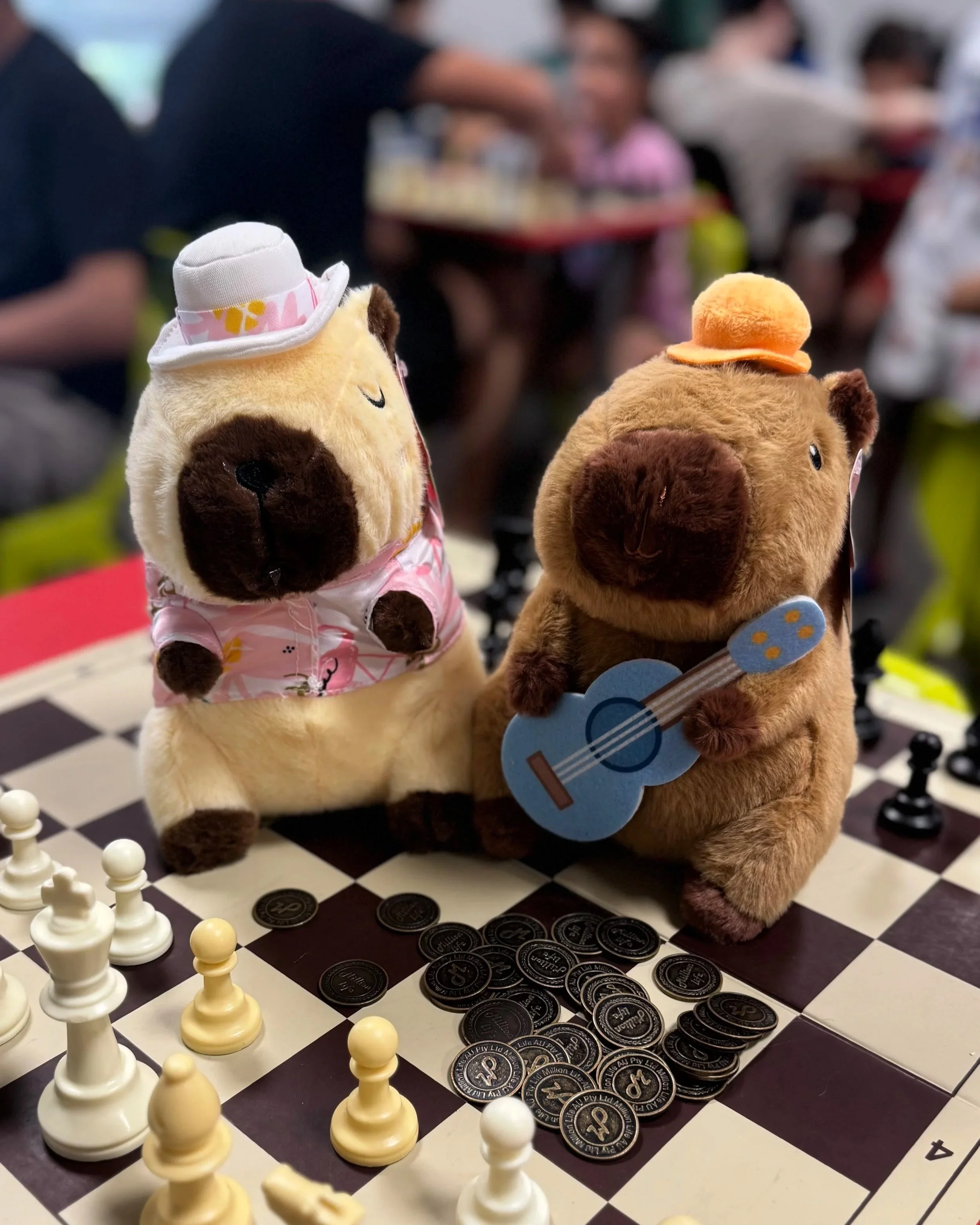 Two plush toy hamsters wearing hats and clothes, one with a small guitar, sit on a chessboard with chess pieces and coins, while people are seen in the background.