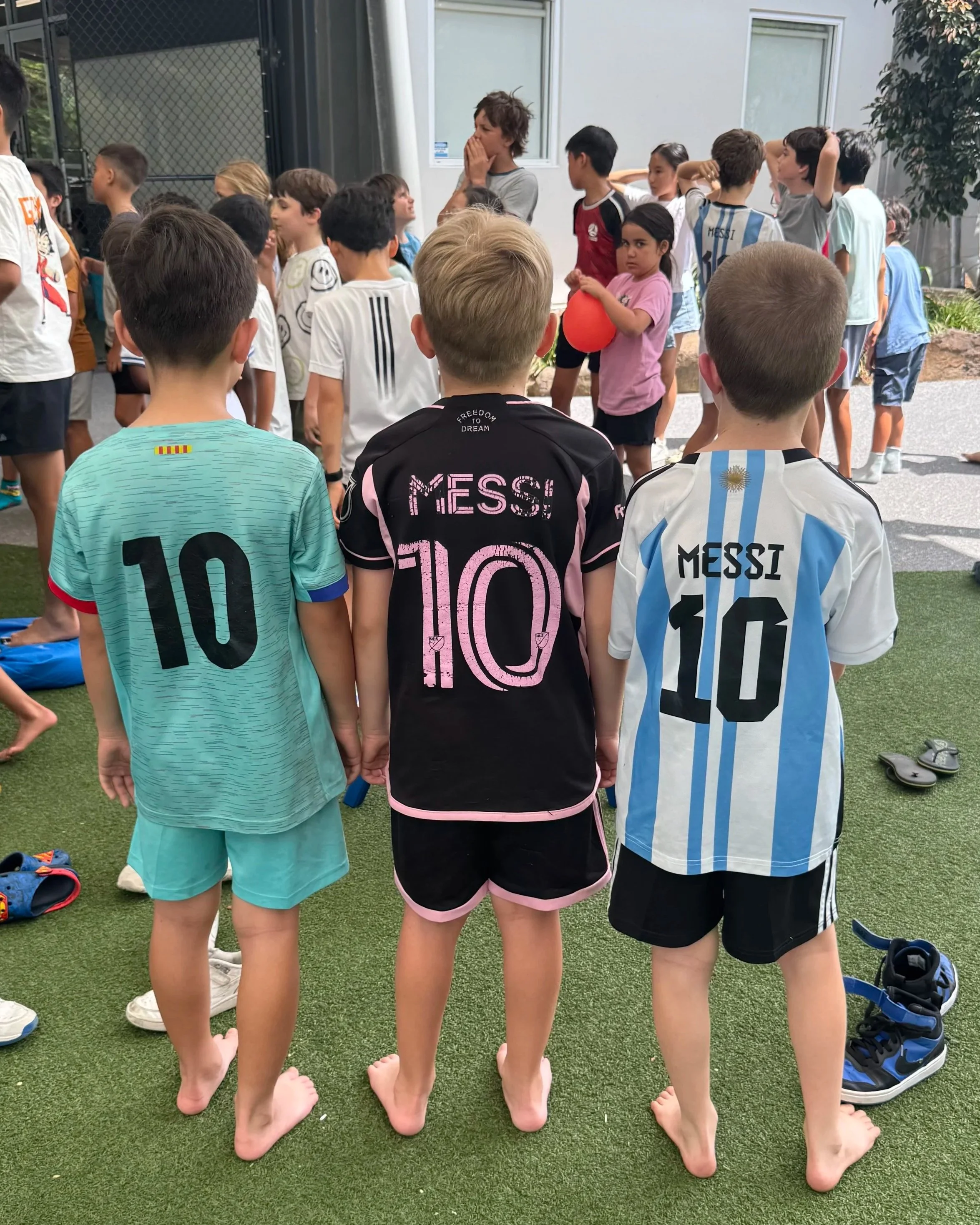 Three children standing on the grass, holding hands, with their backs to the camera, wearing soccer jerseys. There are other children and adults in the background, some talking and some standing in line.