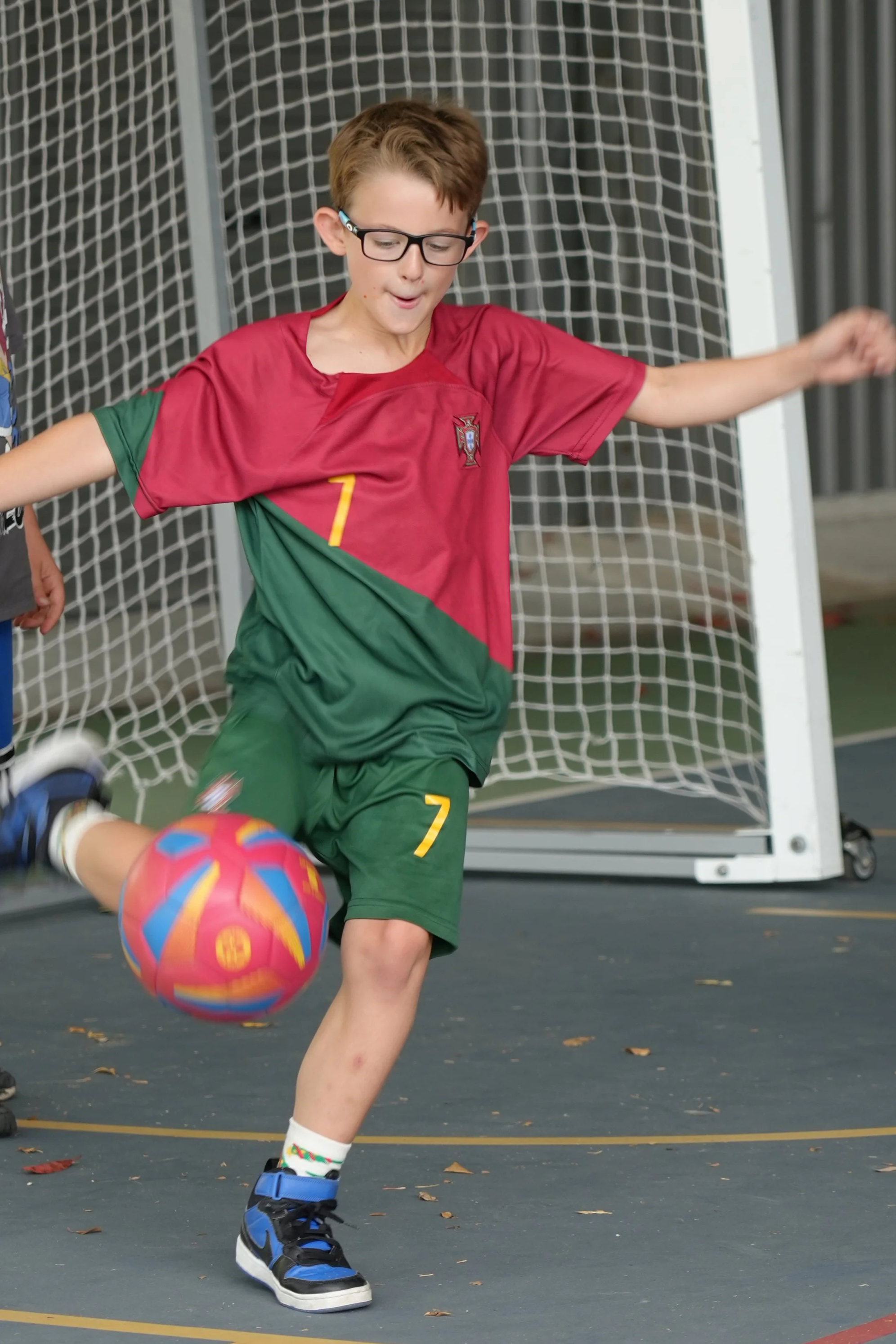 A young boy wearing glasses and a red and green sports uniform kicking a colorful soccer ball on an indoor court in front of a goal net.