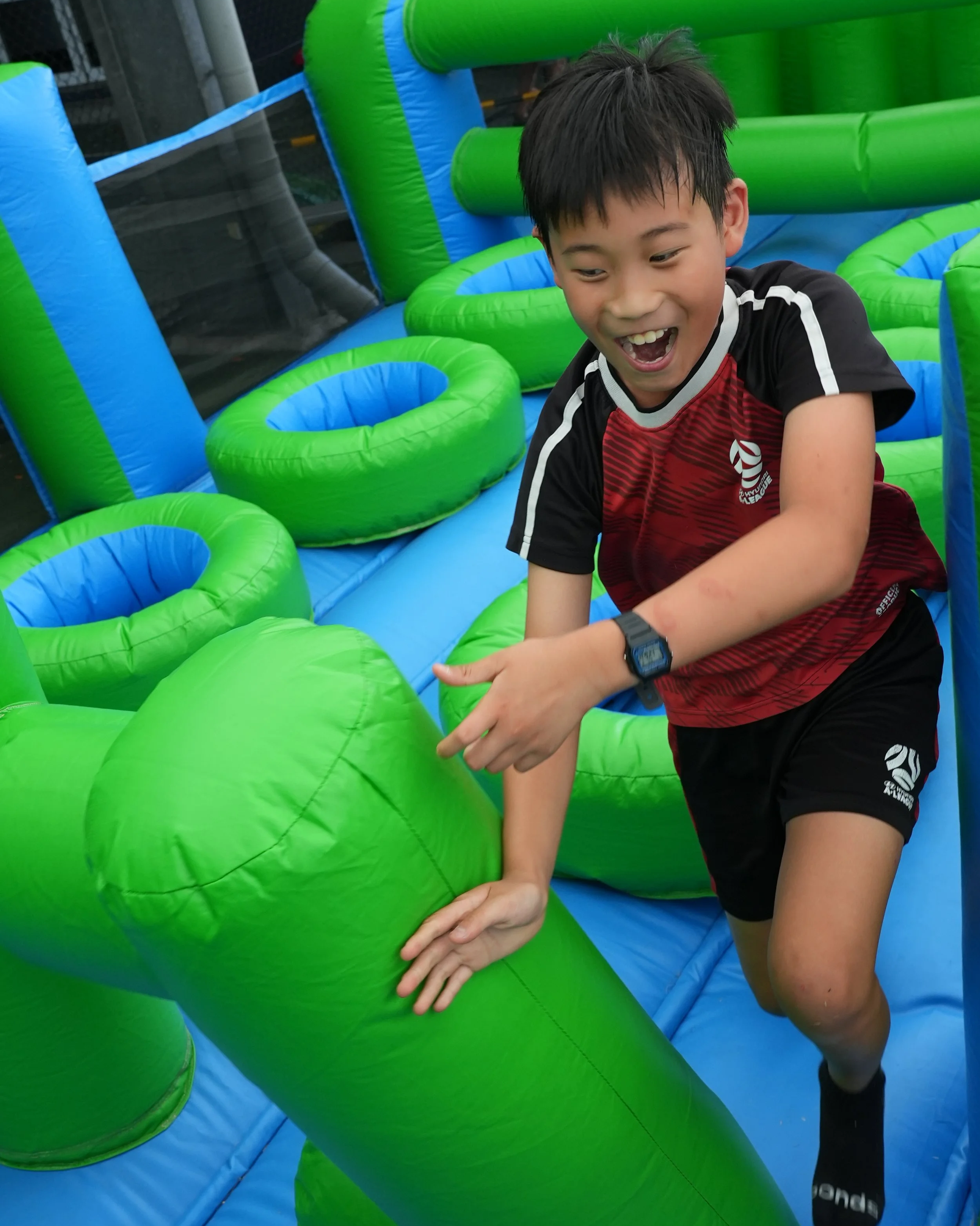 A boy in a black and red athletic shirt and shorts, wearing a black watch, is playing on a colorful inflatable obstacle course. He is holding a large green inflatable object with a smile on his face.