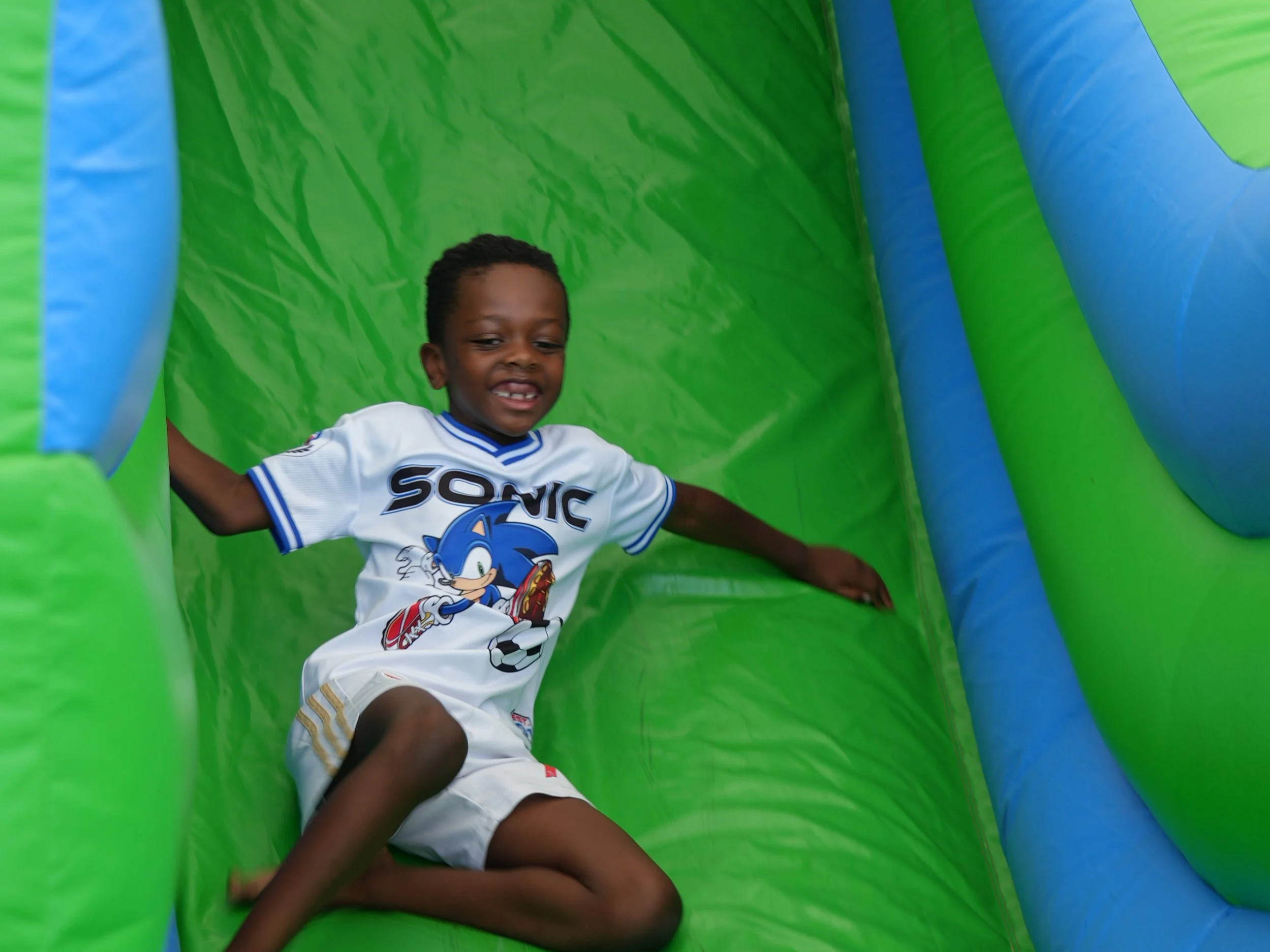 A young boy in a Sonic the Hedgehog t-shirt sliding down a colorful inflatable slide at a play area.