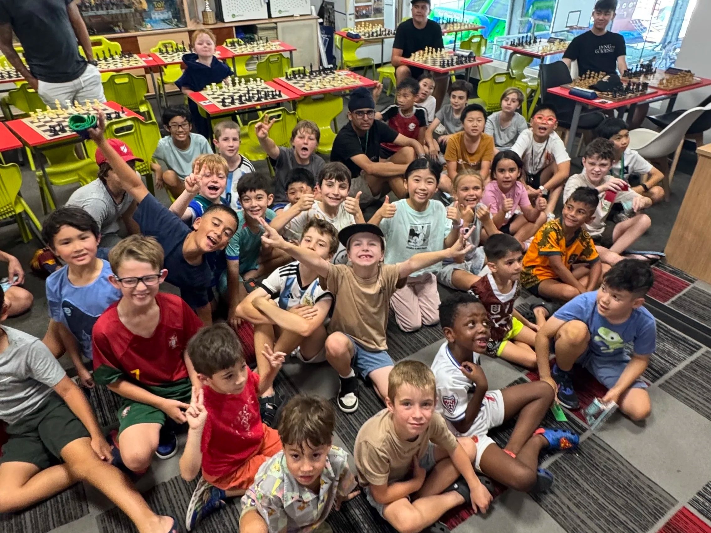 A group of children sitting on a colorful carpet in a chess club or classroom, with some kids making peace signs, smiling, and posing for the camera. In the background, some adults are sitting at tables with chessboards, and a large window reveals an