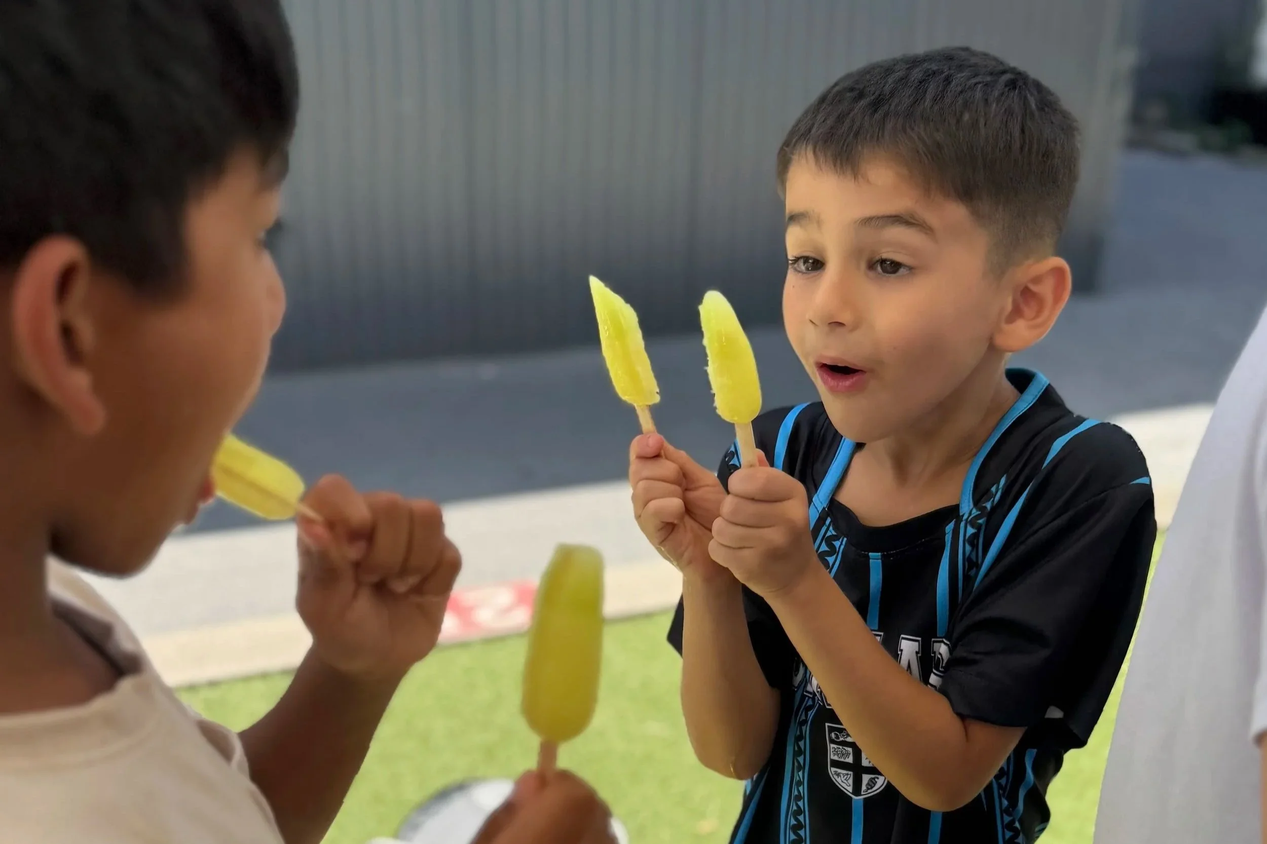 Three young boys standing outside on grass, holding yellow popsicles, engaging in conversation.