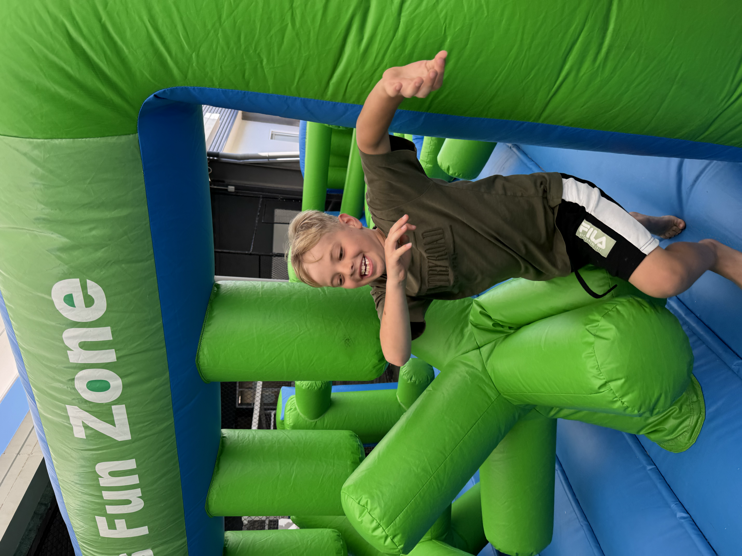 A young boy is playing inside a green and blue inflatable bounce house, smiling and making a peace sign with his hand.