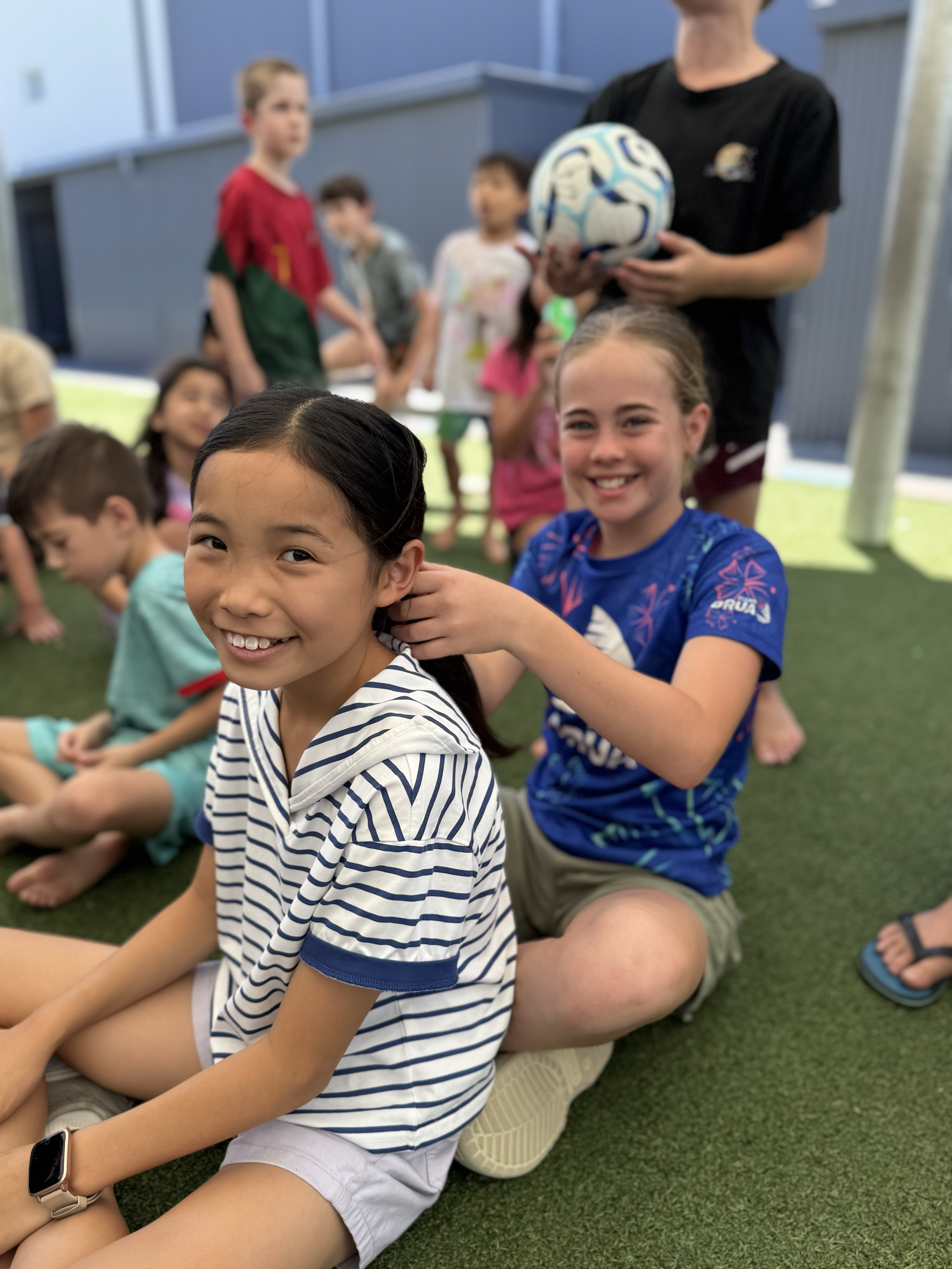 Smiling girl in a striped shirt sitting on the ground while another girl in a blue soccer jersey adjusts her hair. Children are sitting and standing on a grassy area with a blue building in the background.