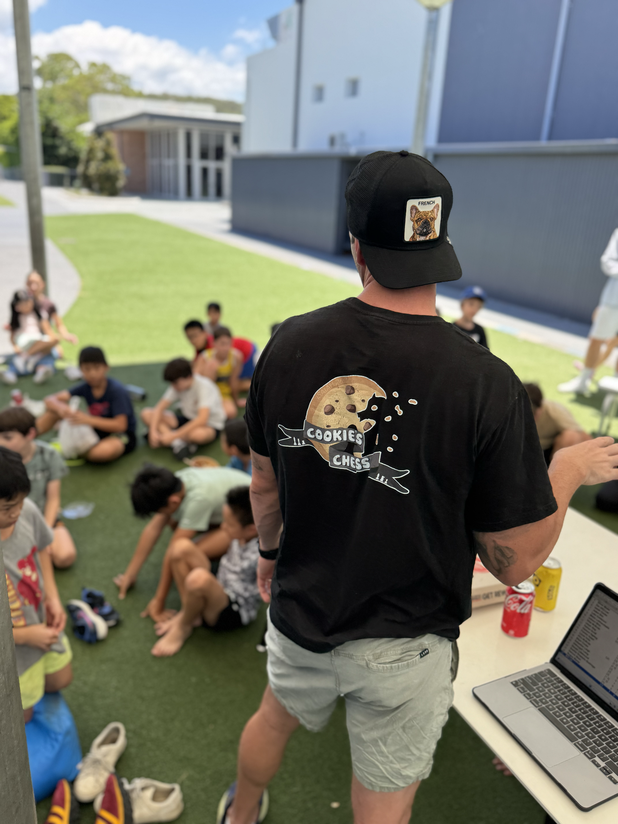 A man wearing a black hat with a French Bulldog patch and a black t-shirt with a cookie design and the text 'Cookies and Chess' stands in front of a group of children sitting on artificial grass outdoors, with a table, laptops, and drinks nearby.