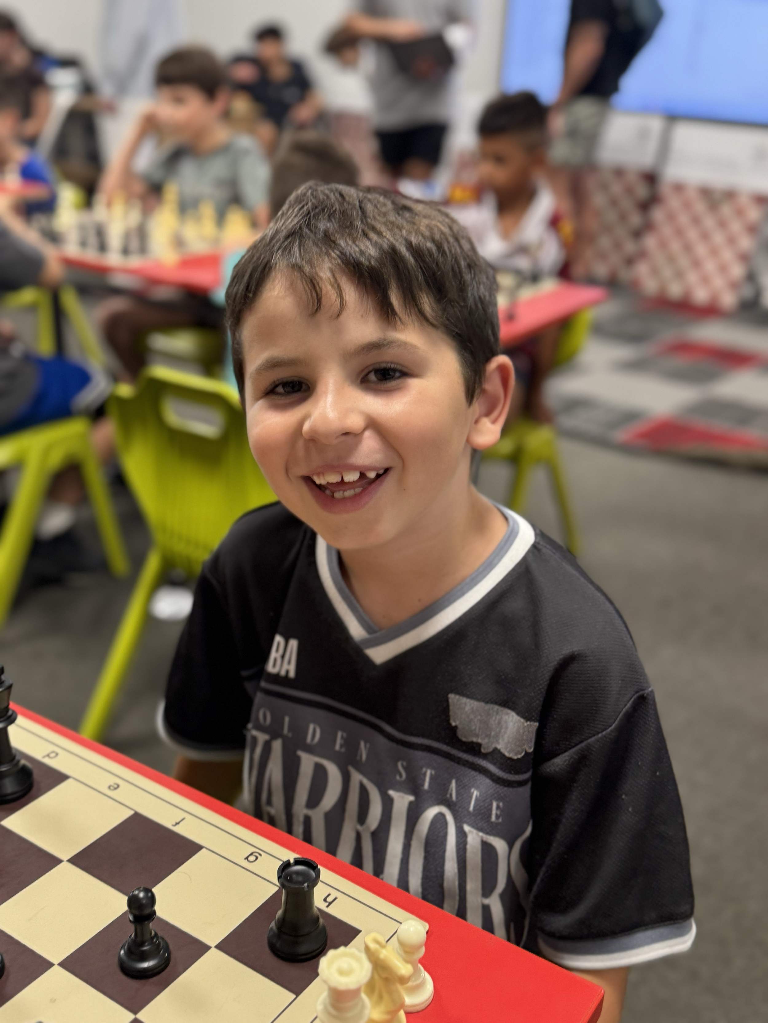 Young boy smiling at the camera while playing chess with chess pieces on a red table in a busy classroom with other children and adults in the background.