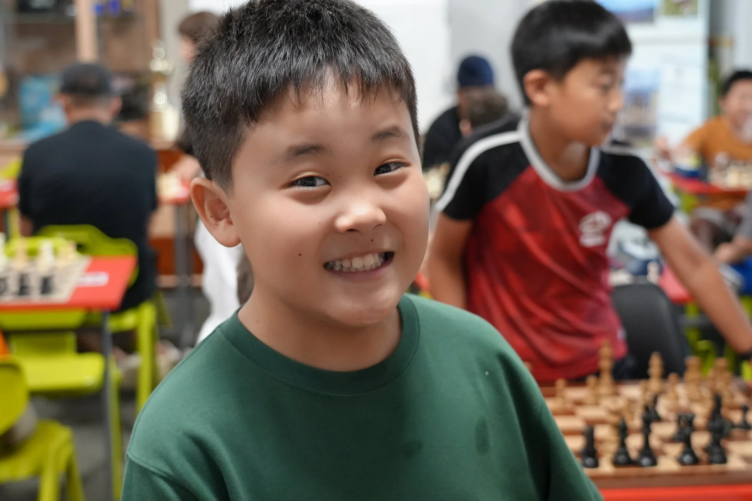 Young boy smiling at a chess tournament in a crowded room, other children and adults in the background.