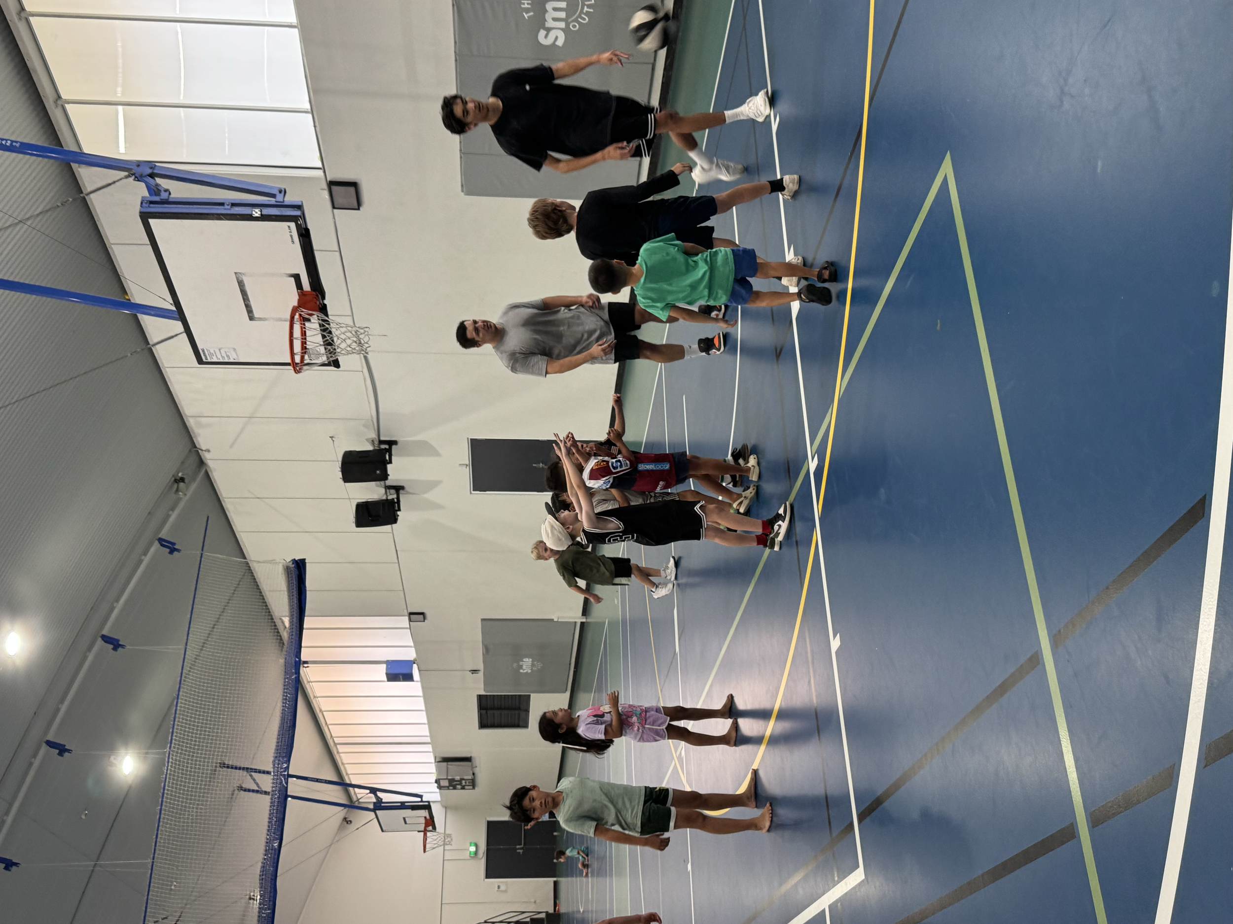 Group of children and adults standing on an indoor basketball court, some talking and others stretching. There are two basketball hoops, one at each end of the court. The floor is blue with white, yellow, and black lines. The walls are white with a b