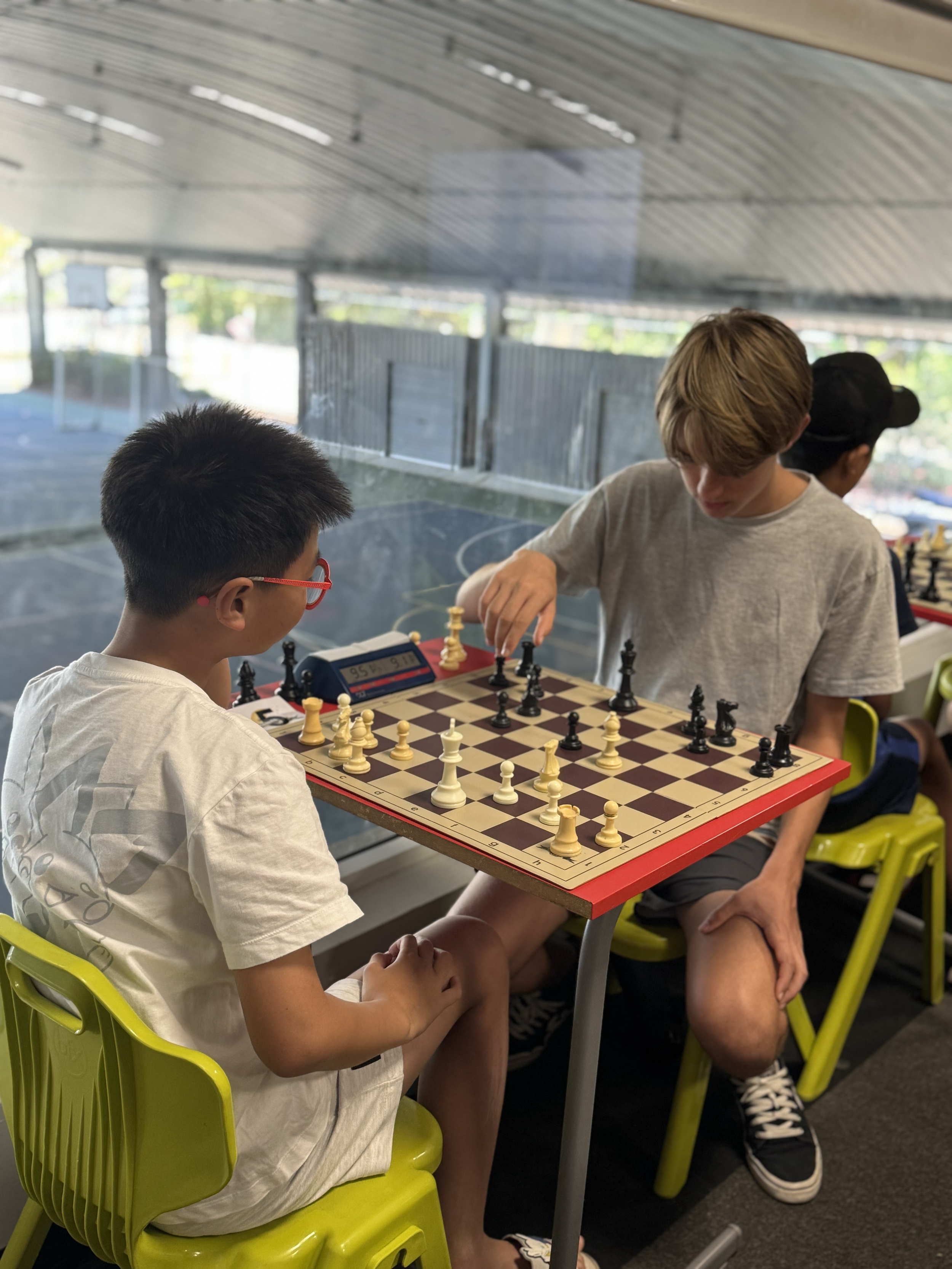 Two young boys playing chess at a small table in an indoor area near a large window, with a view of an outdoor sports court.